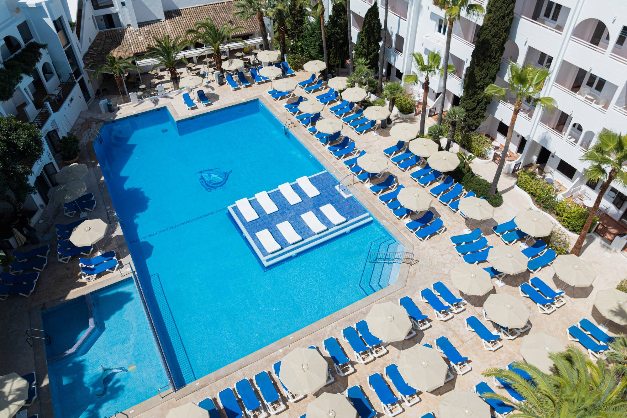 Resort swimming pool with blue loungers, umbrellas, white buildings, and palm trees.
