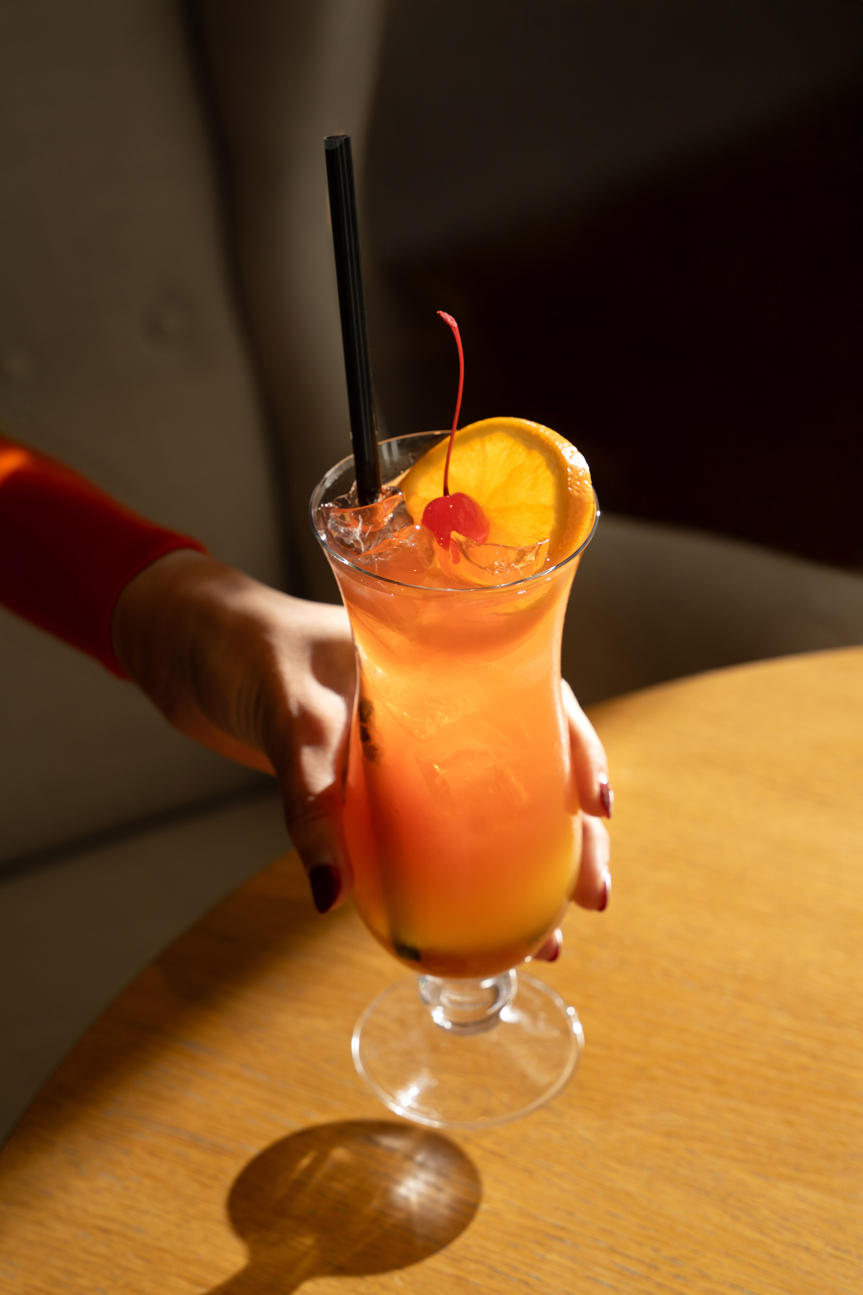 Orange cocktail with cherry, orange slice, and black straw on a wooden table.