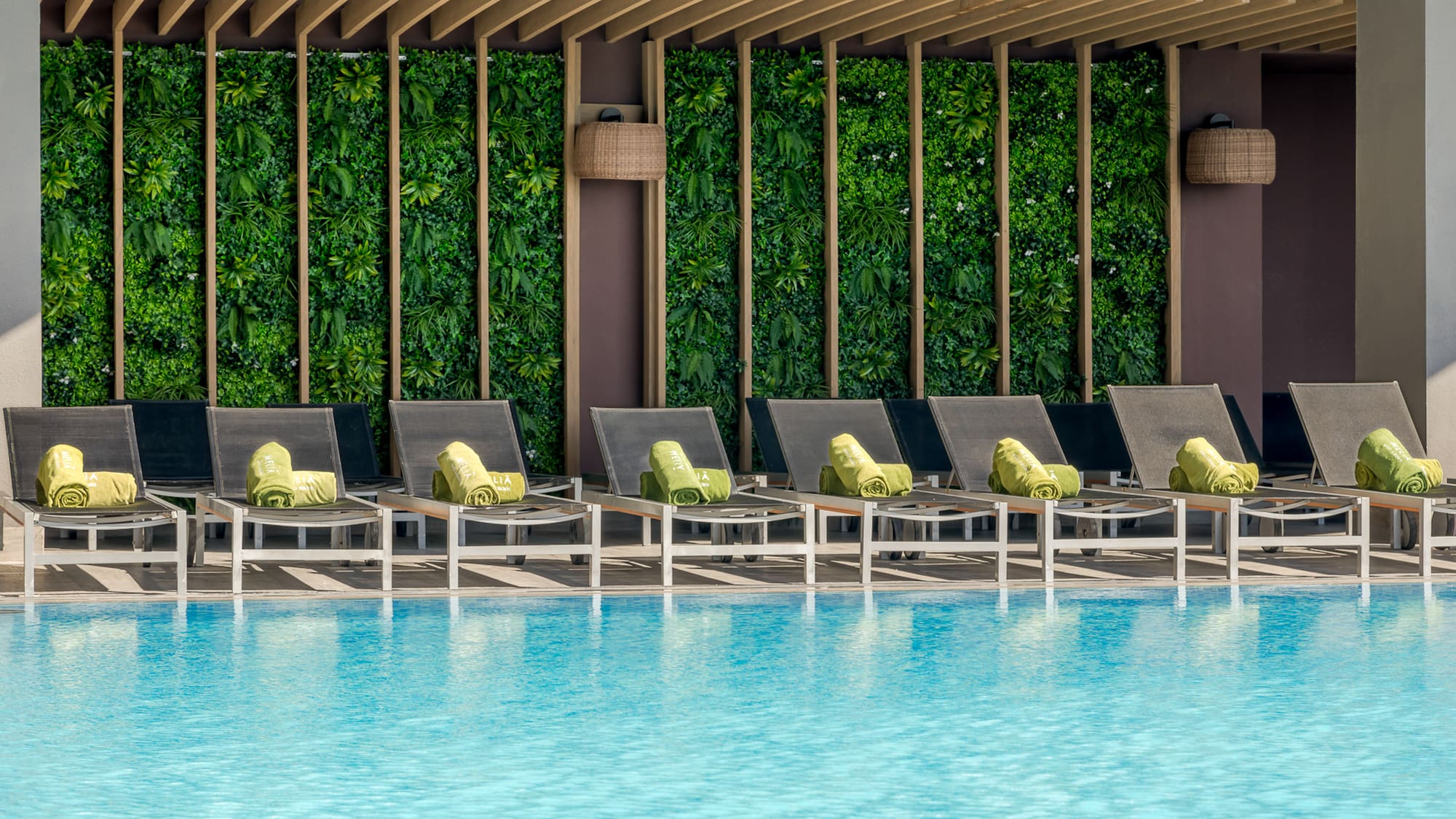 Piscina de hotel con tumbonas, toallas verdes y jardín vertical.