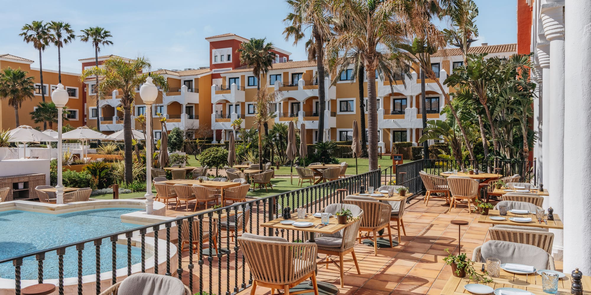 Outdoor resort restaurant terrace with set tables, palm trees, and pool.