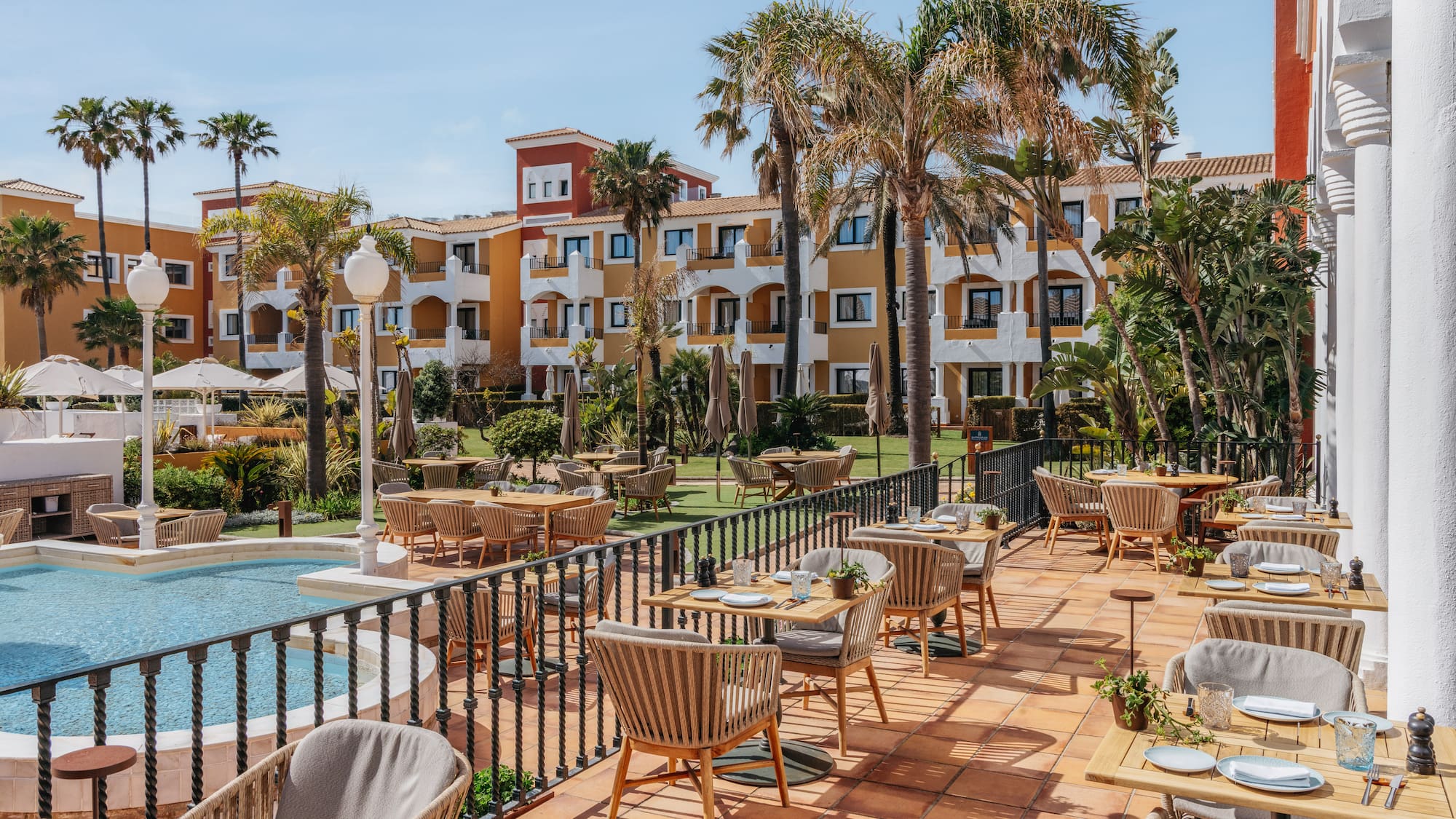 Sunny resort outdoor dining terrace with palm trees, pool, and hotel buildings.