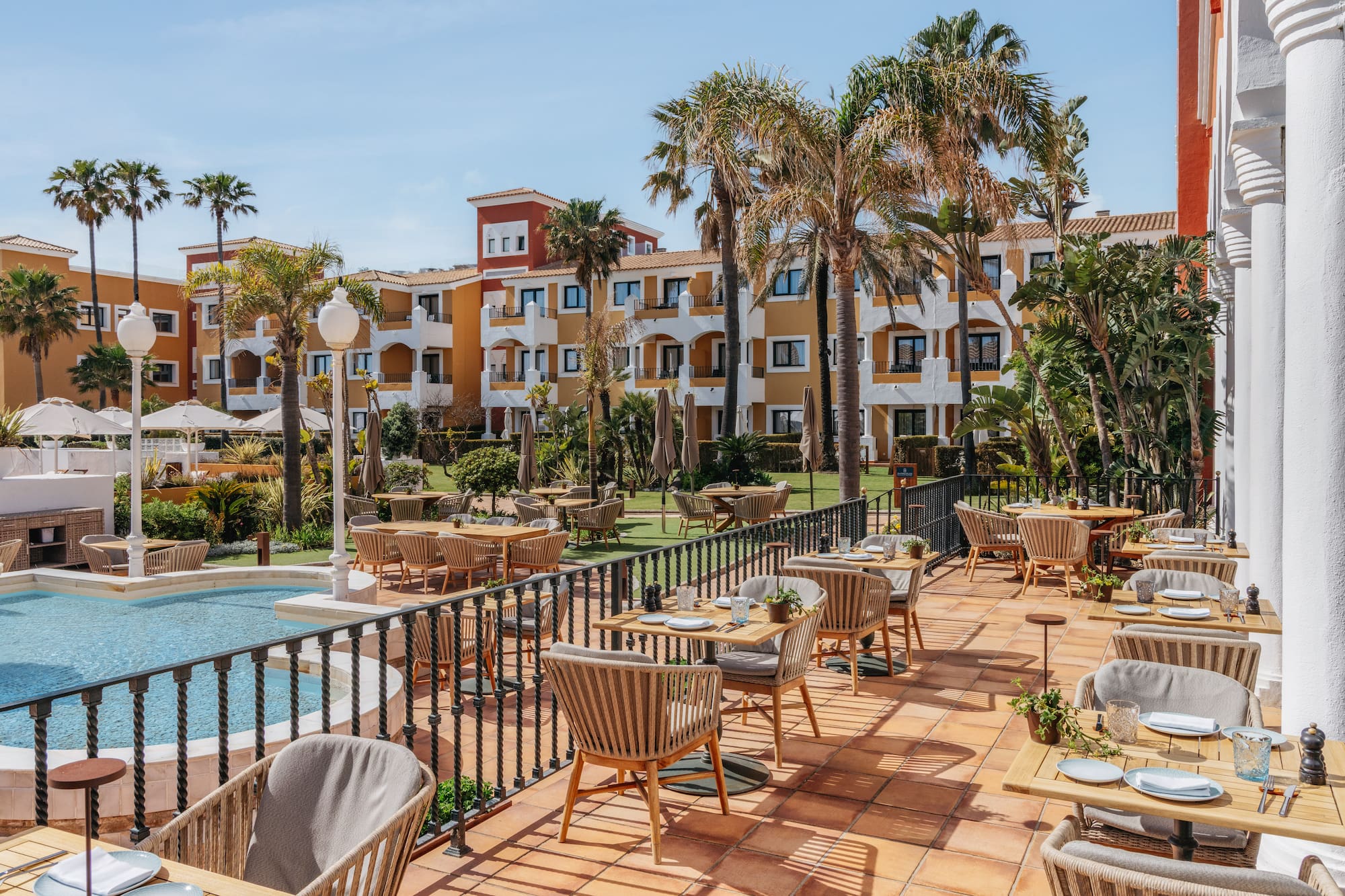 Outdoor resort restaurant terrace with set tables, palm trees, and pool.