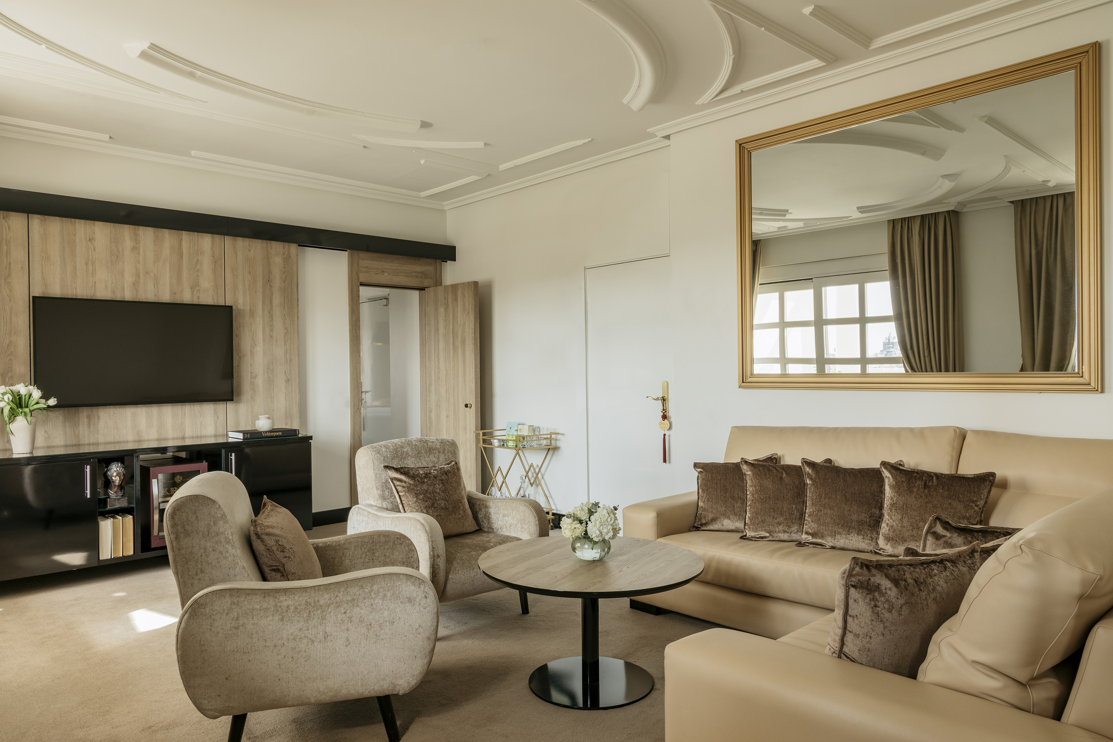 Elegant hotel suite living room with beige sofa, armchairs, TV, and mirror.