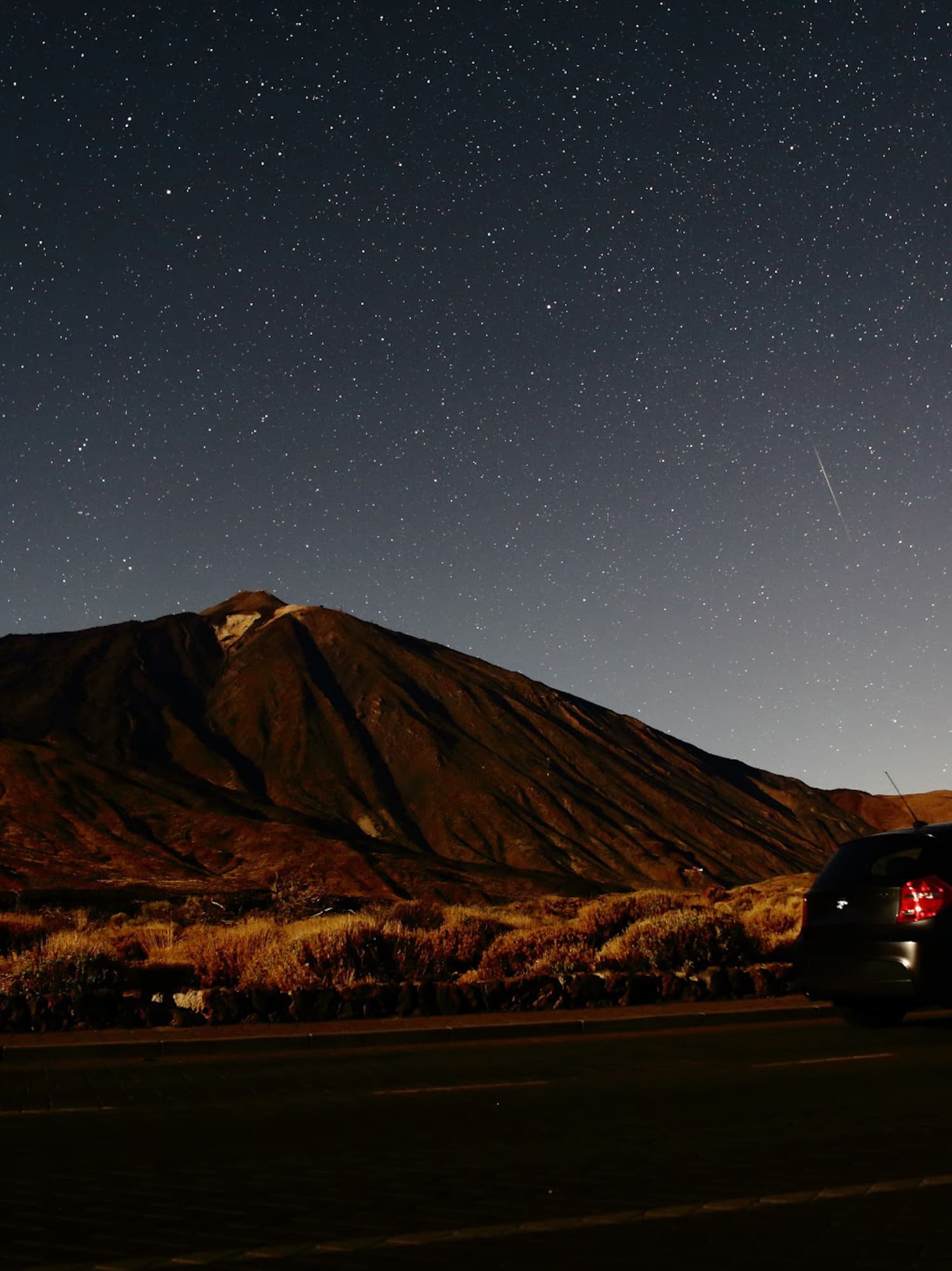 Dark car on road under vast starry night sky, large volcanic mountain in background.