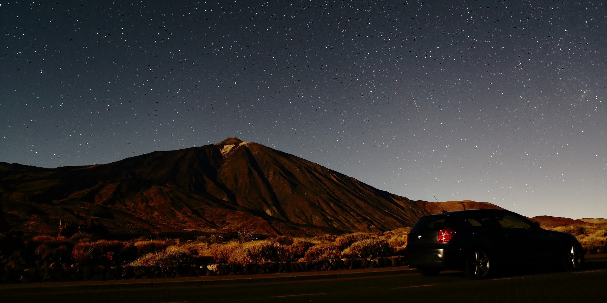 Dark car on road under vast starry night sky, large volcanic mountain in background.