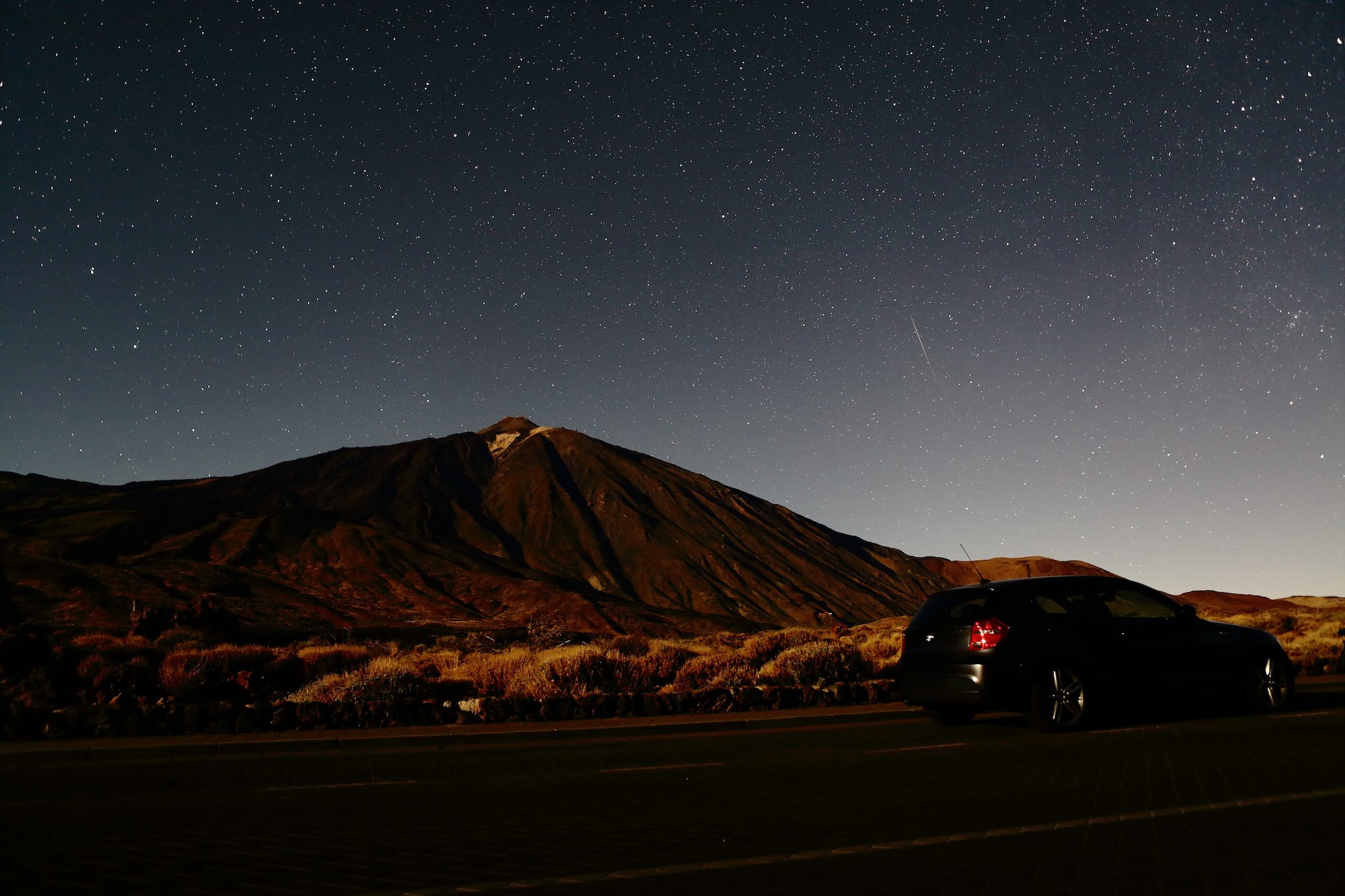 Dark car on road under vast starry night sky, large volcanic mountain in background.