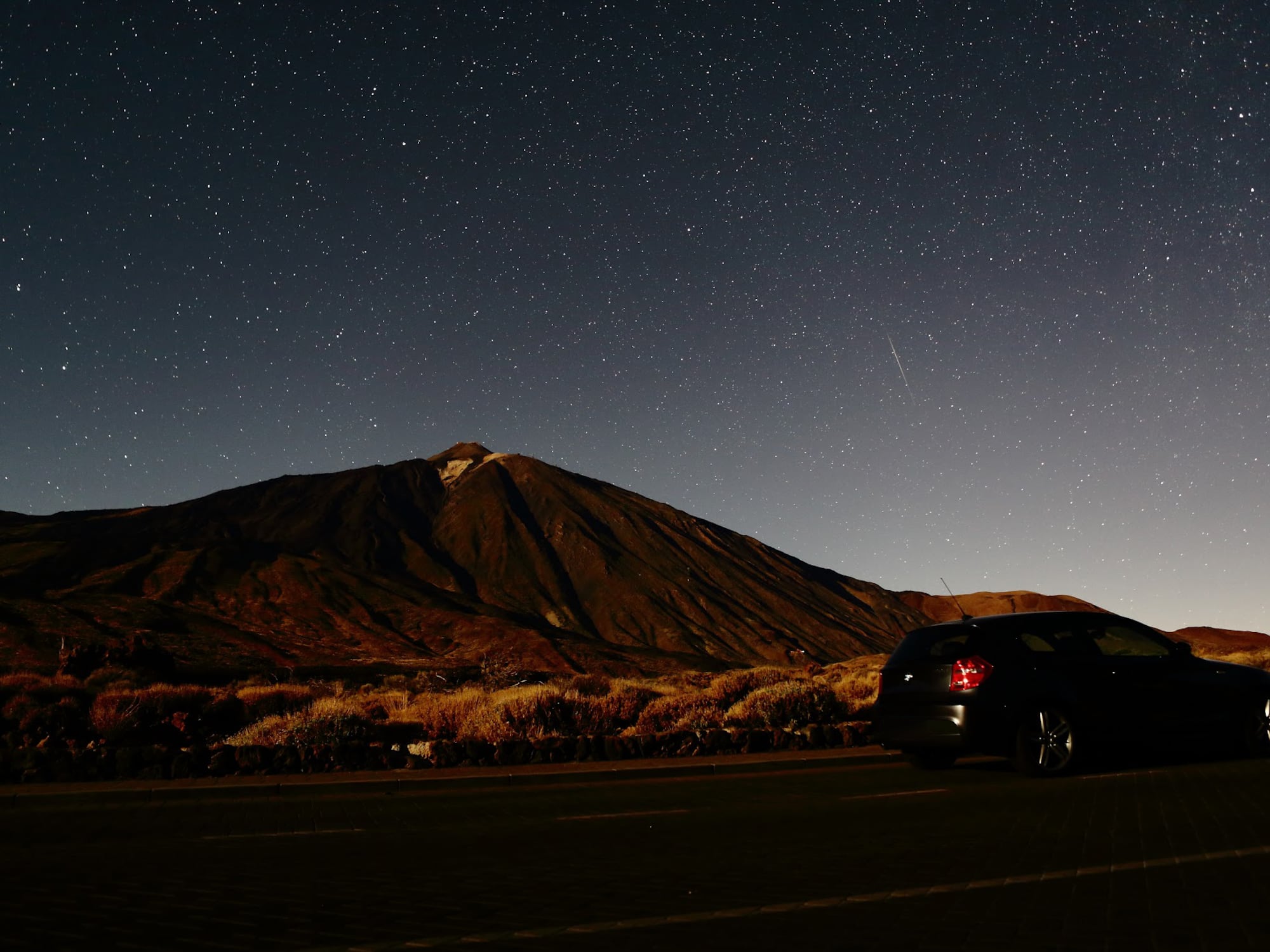 Dark car on road under vast starry night sky, large volcanic mountain in background.