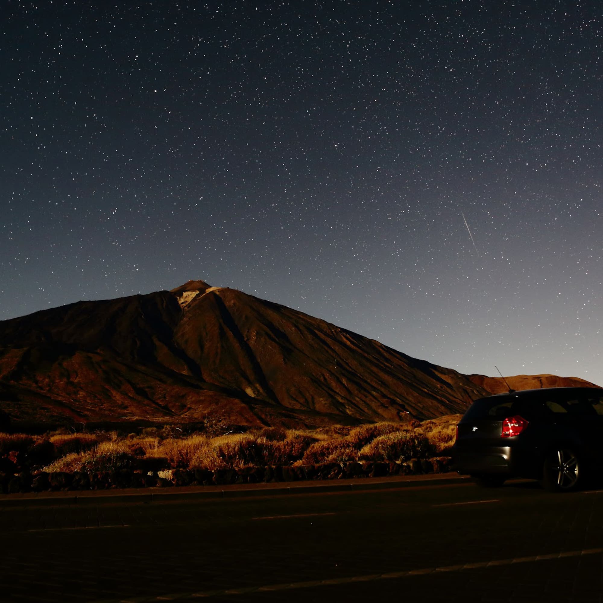 Dark car on road under vast starry night sky, large volcanic mountain in background.