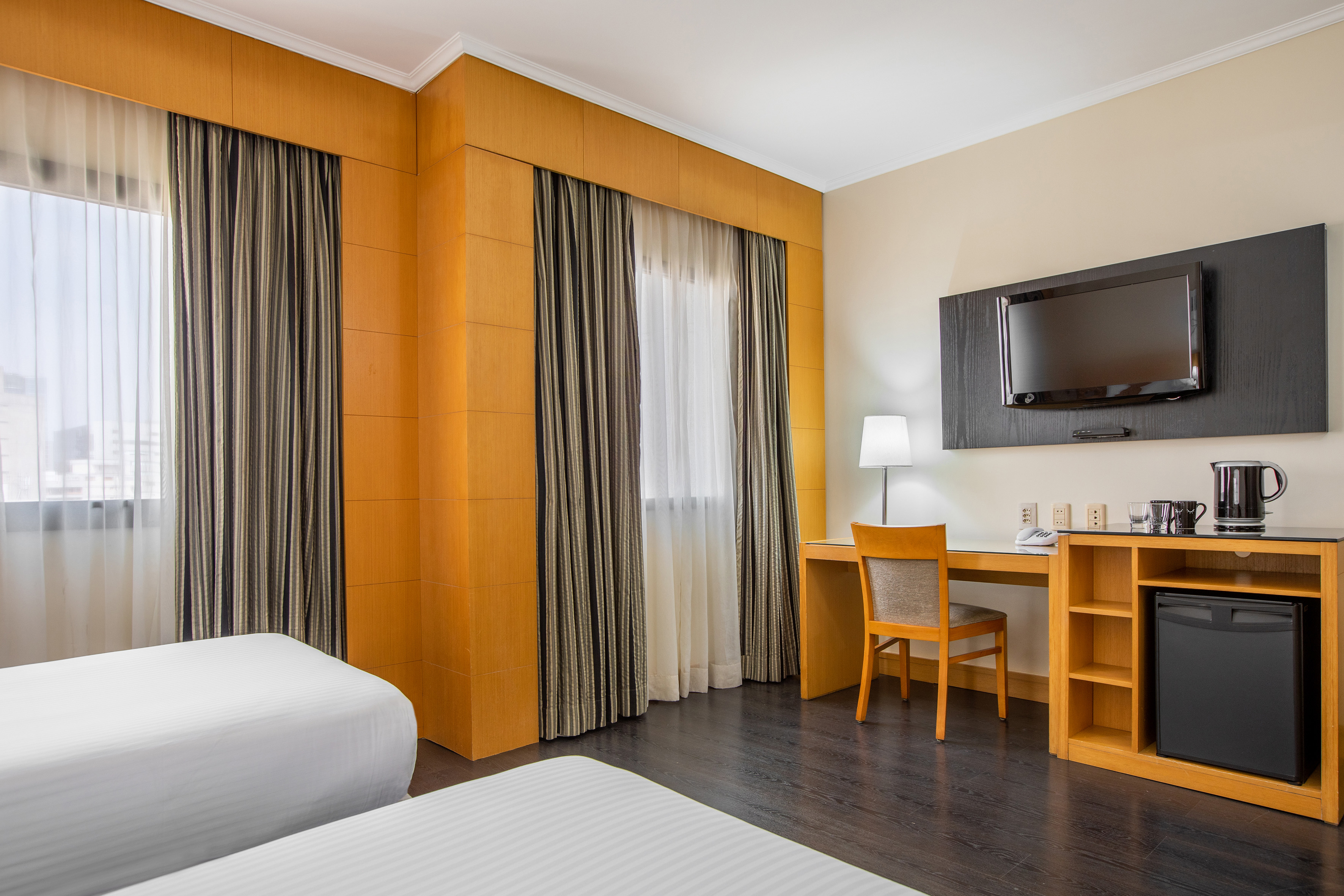 Modern hotel room with twin beds, work desk, wall-mounted TV, and mini-bar.