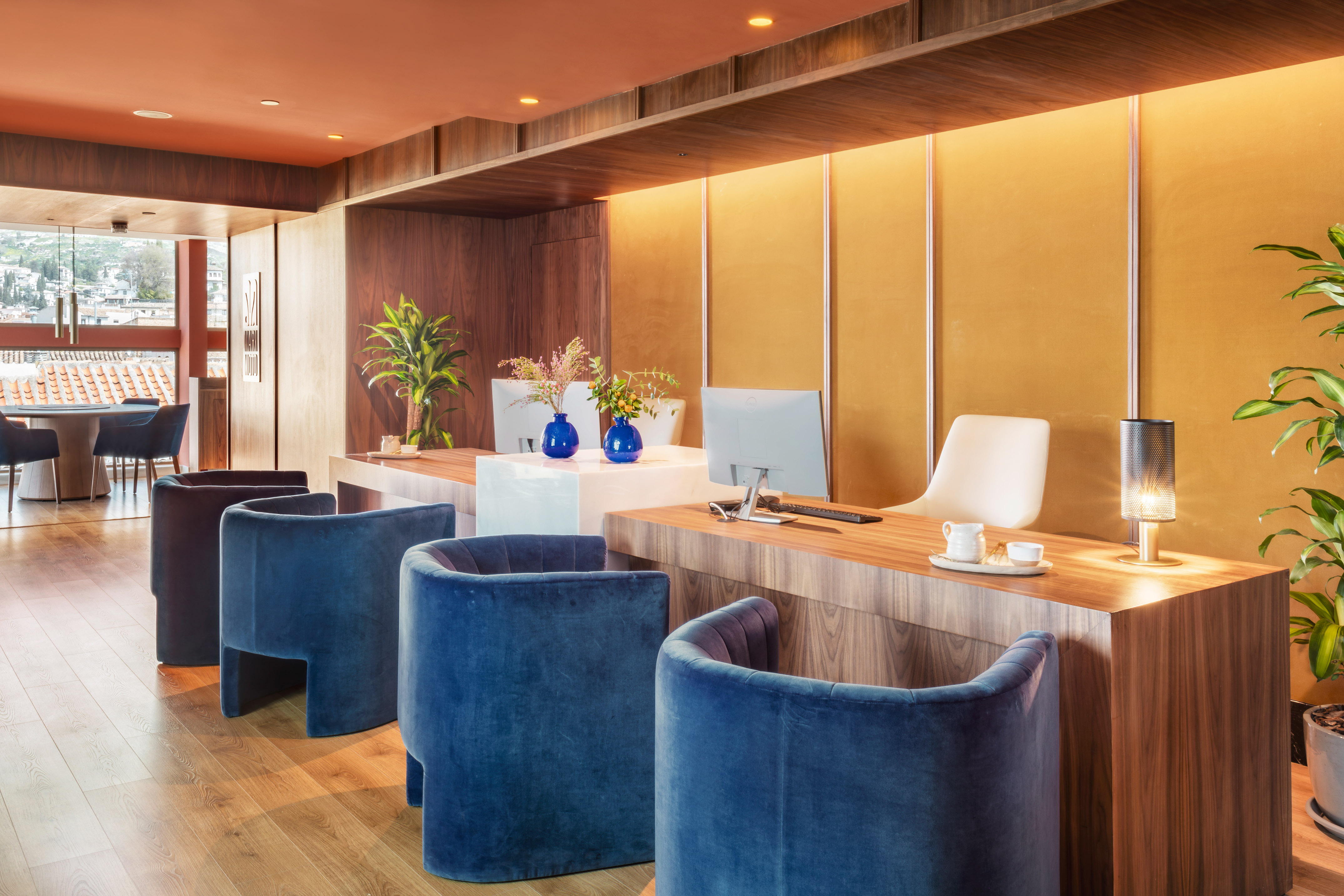 Modern hotel reception with wooden desk, blue velvet chairs, and city view.