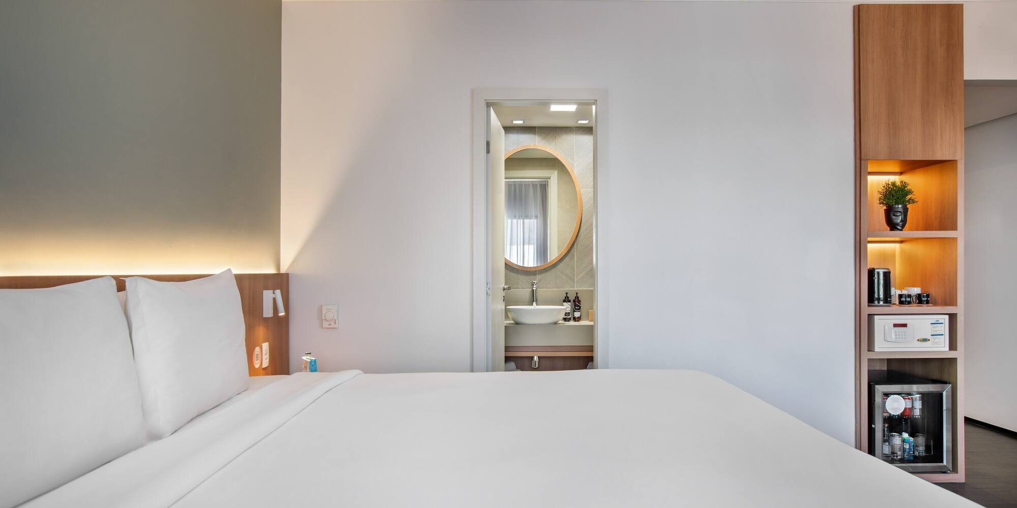 Modern hotel bedroom with white bed, open bathroom, and built-in mini-bar.