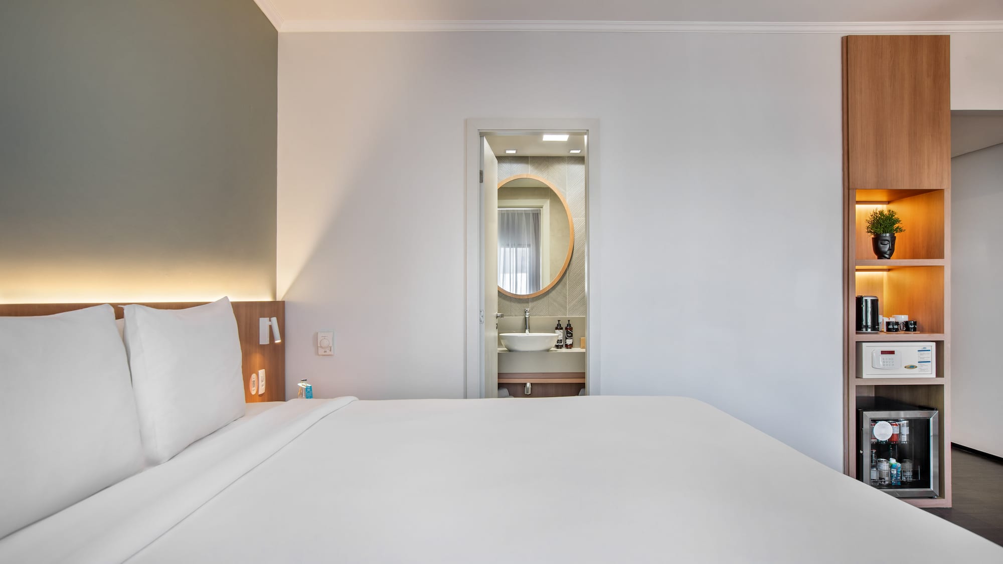 Modern hotel bedroom with white bed, open bathroom, and built-in mini-bar.