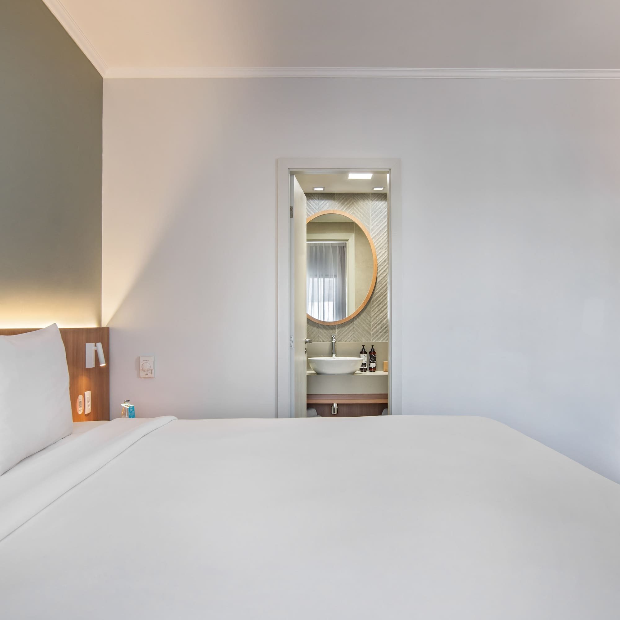 Modern hotel bedroom with white bed, open bathroom, and built-in mini-bar.
