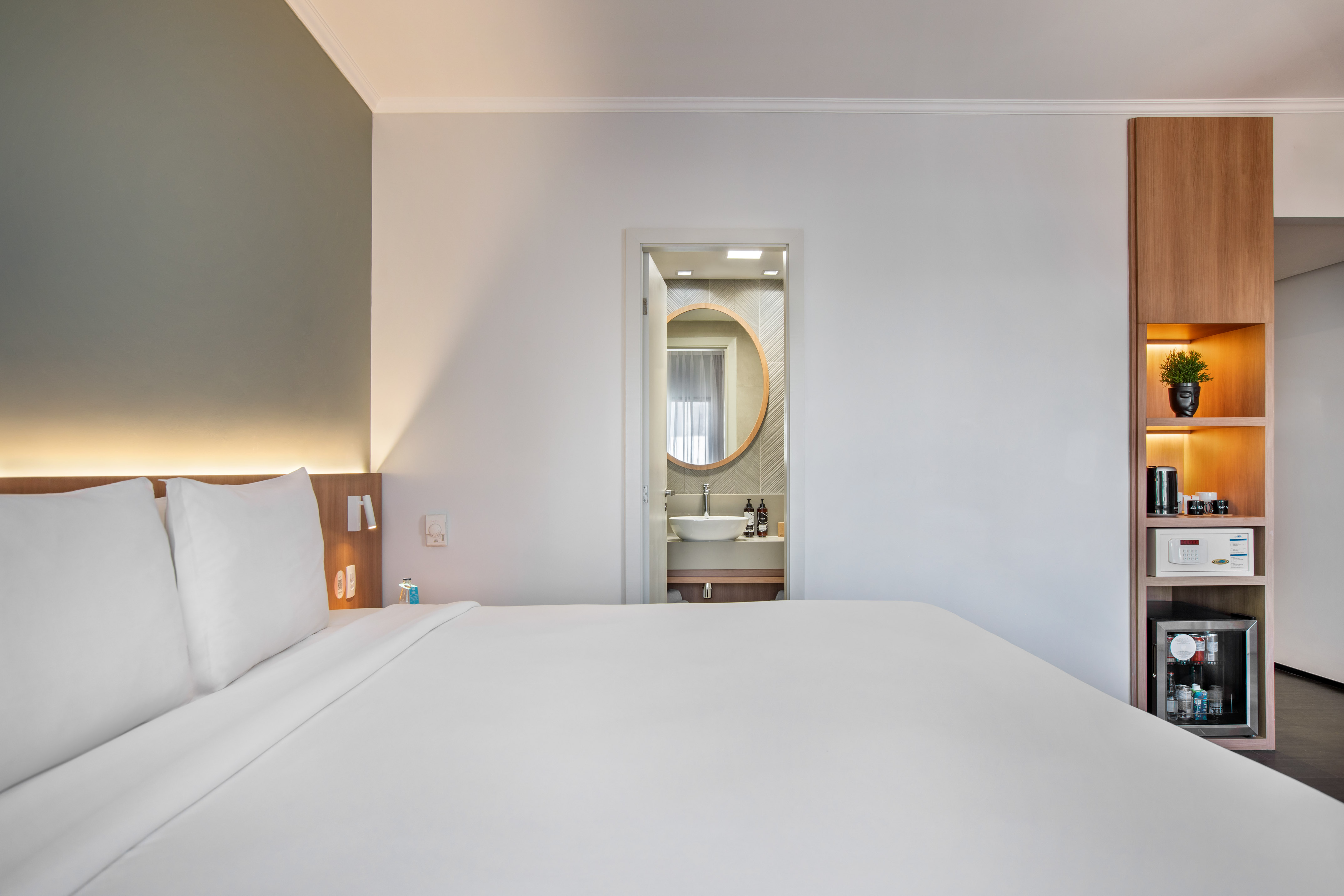 Modern hotel bedroom with white bed, open bathroom, and built-in mini-bar.