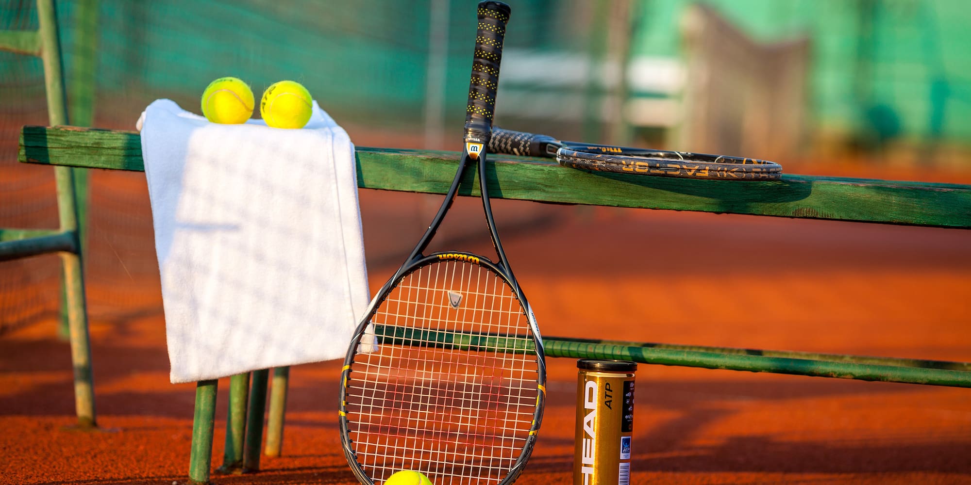 Head tennis racquets, yellow balls, and white towel on a red clay court.