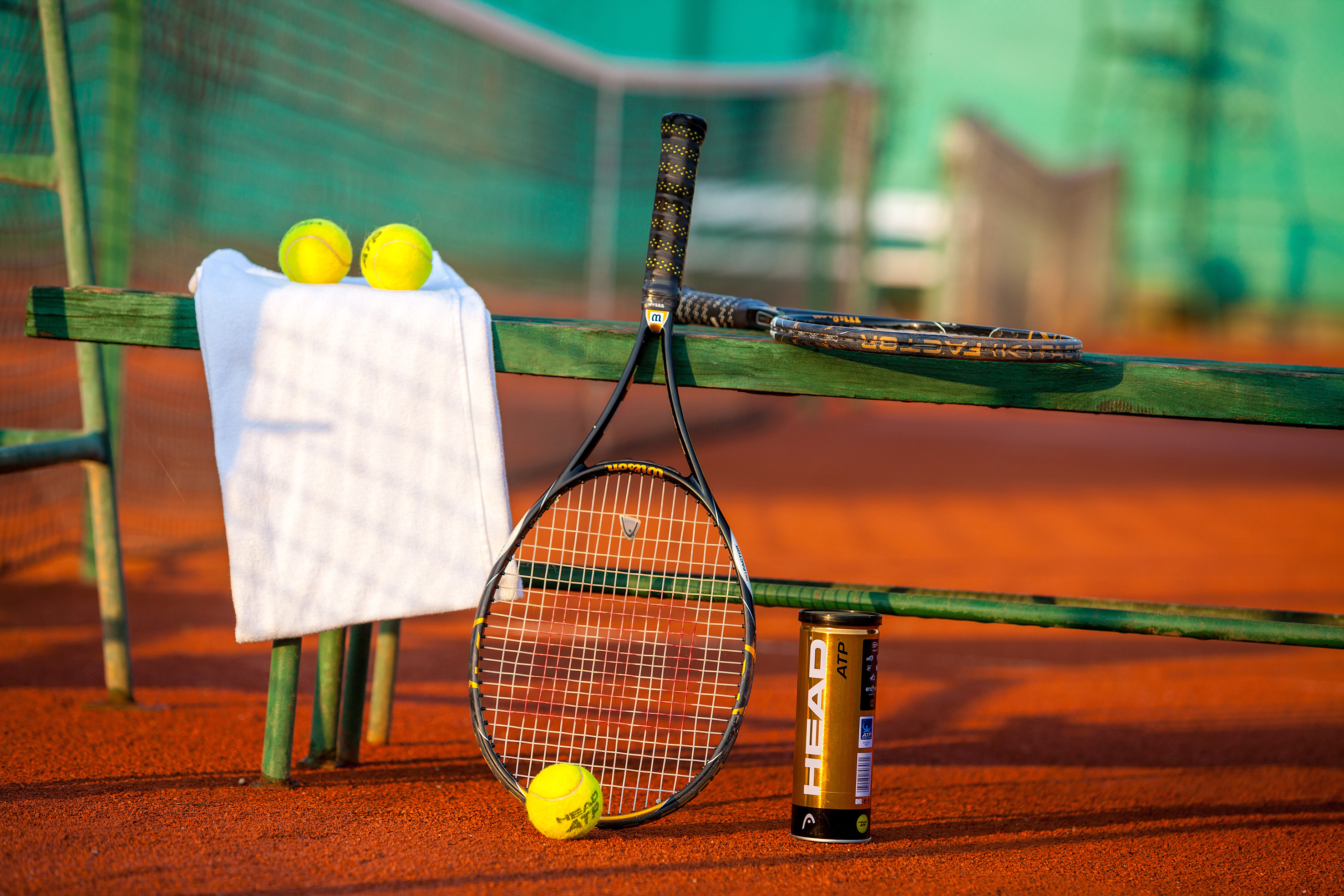 Head tennis racquets, yellow balls, and white towel on a red clay court.
