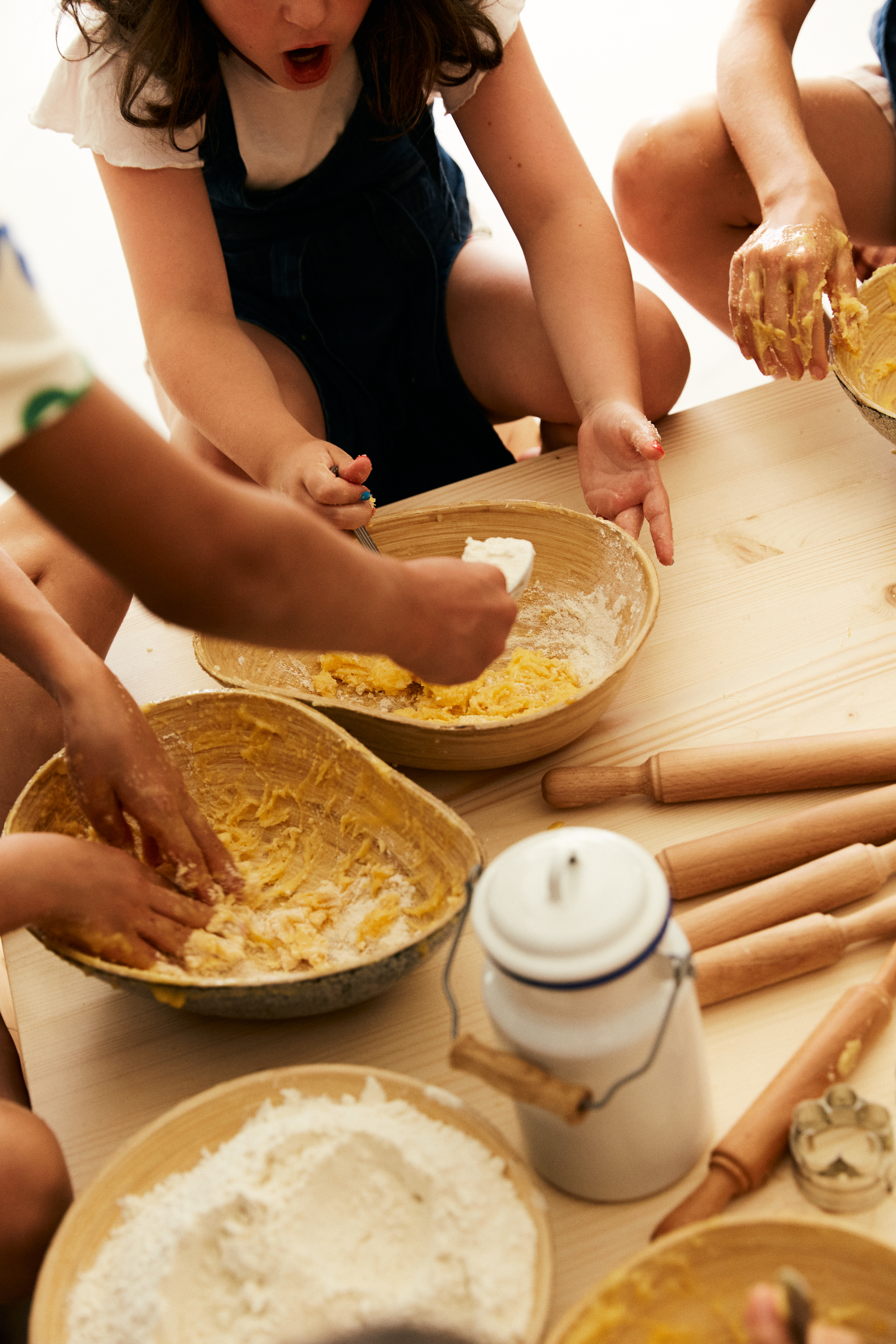 Kids' hands mixing dough in wooden bowls; baking with flour and rolling pins.