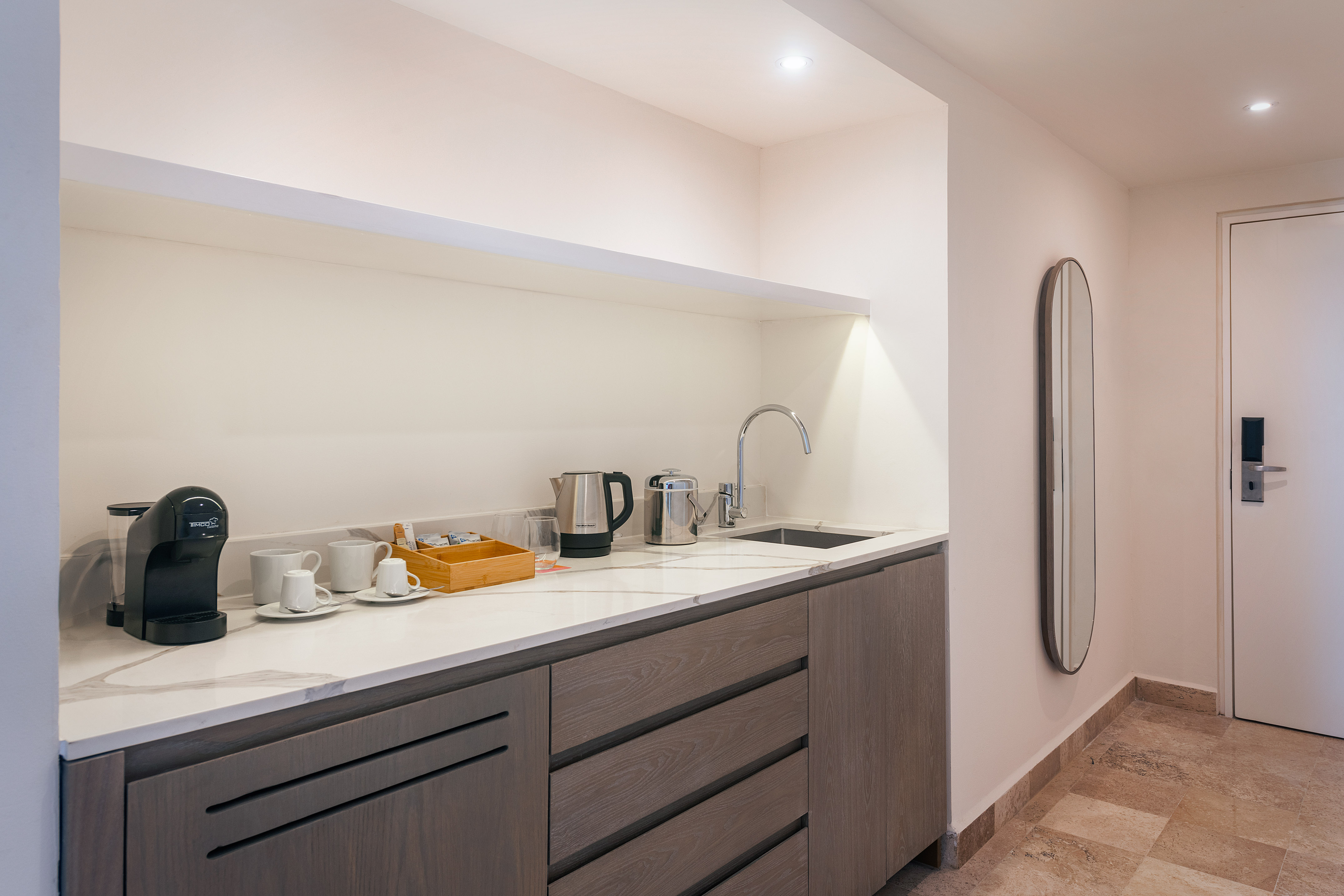 Modern hotel kitchenette with coffee station, sink, kettle, and wooden cabinets.