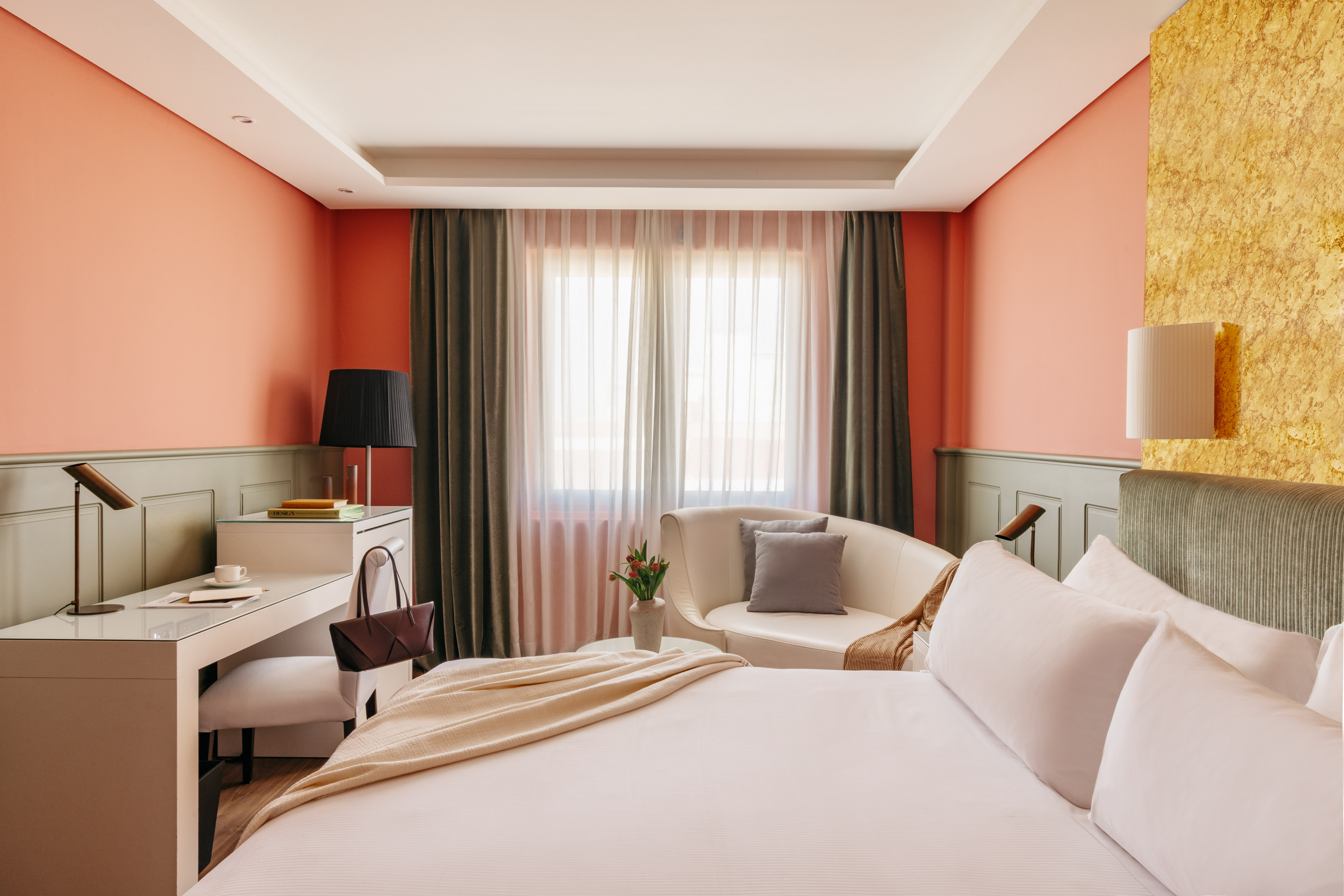 Modern hotel bedroom with coral walls, white bed, desk, and sitting area.