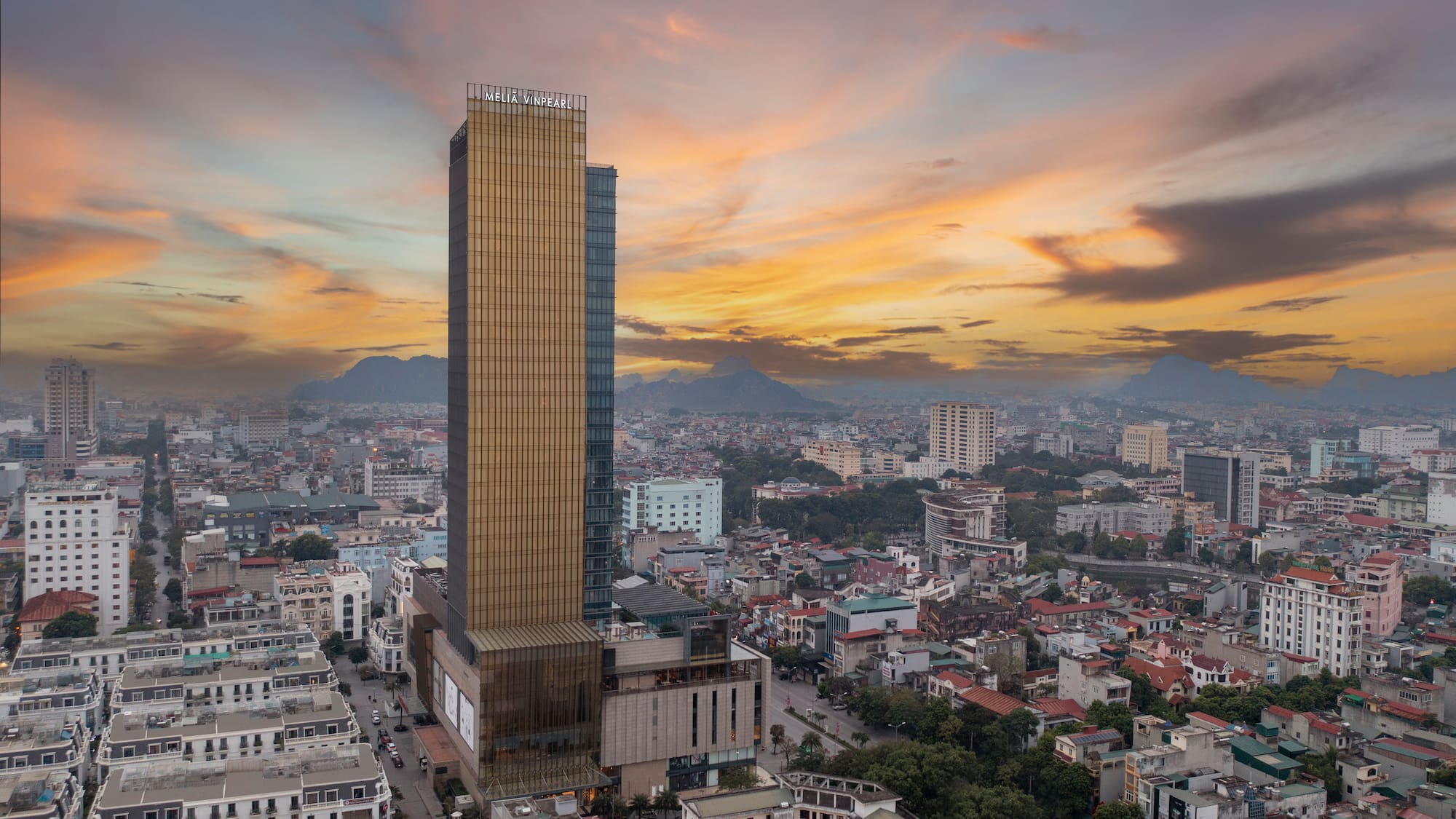 Meliá Vinpearl hotel skyscraper, golden facade, city and mountains at sunset.