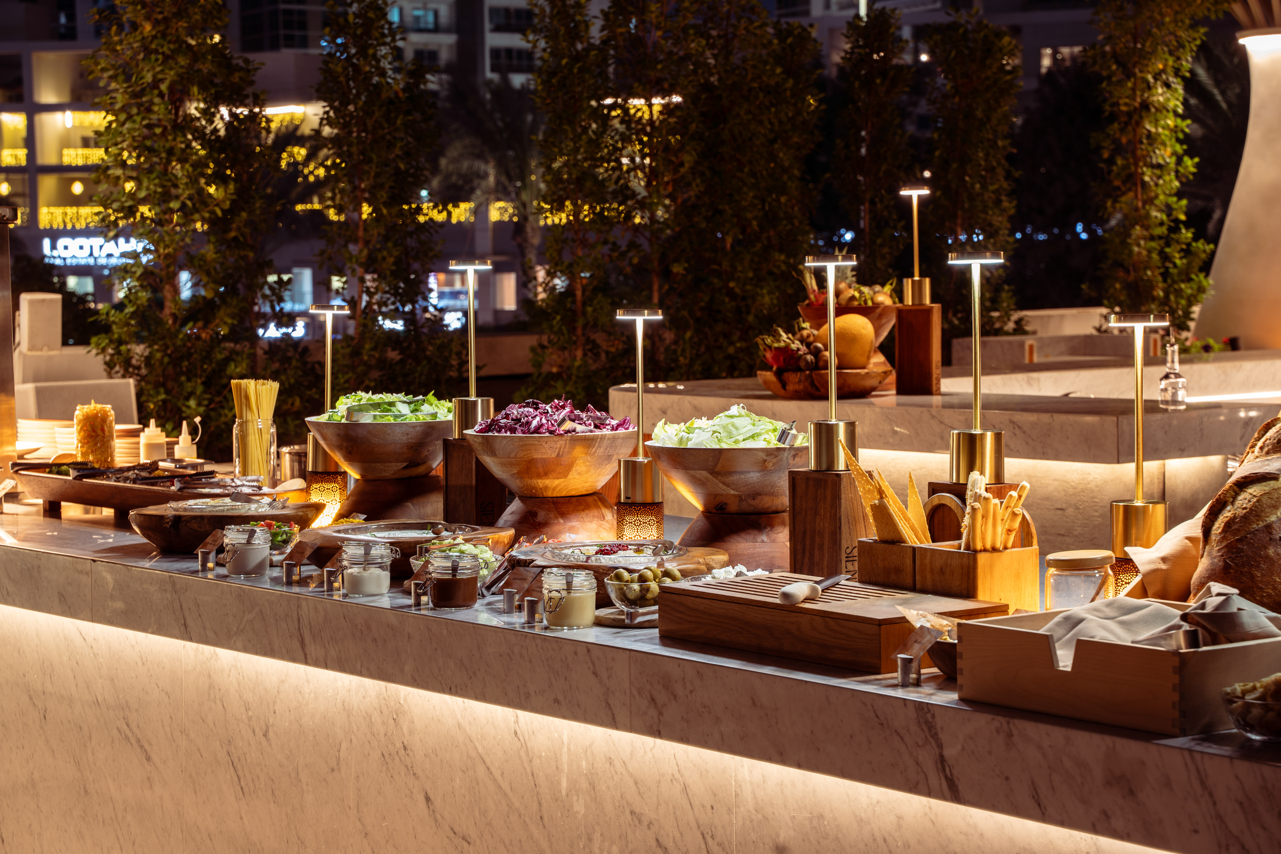 Outdoor evening salad bar with fresh greens, pasta, and dressings on marble.