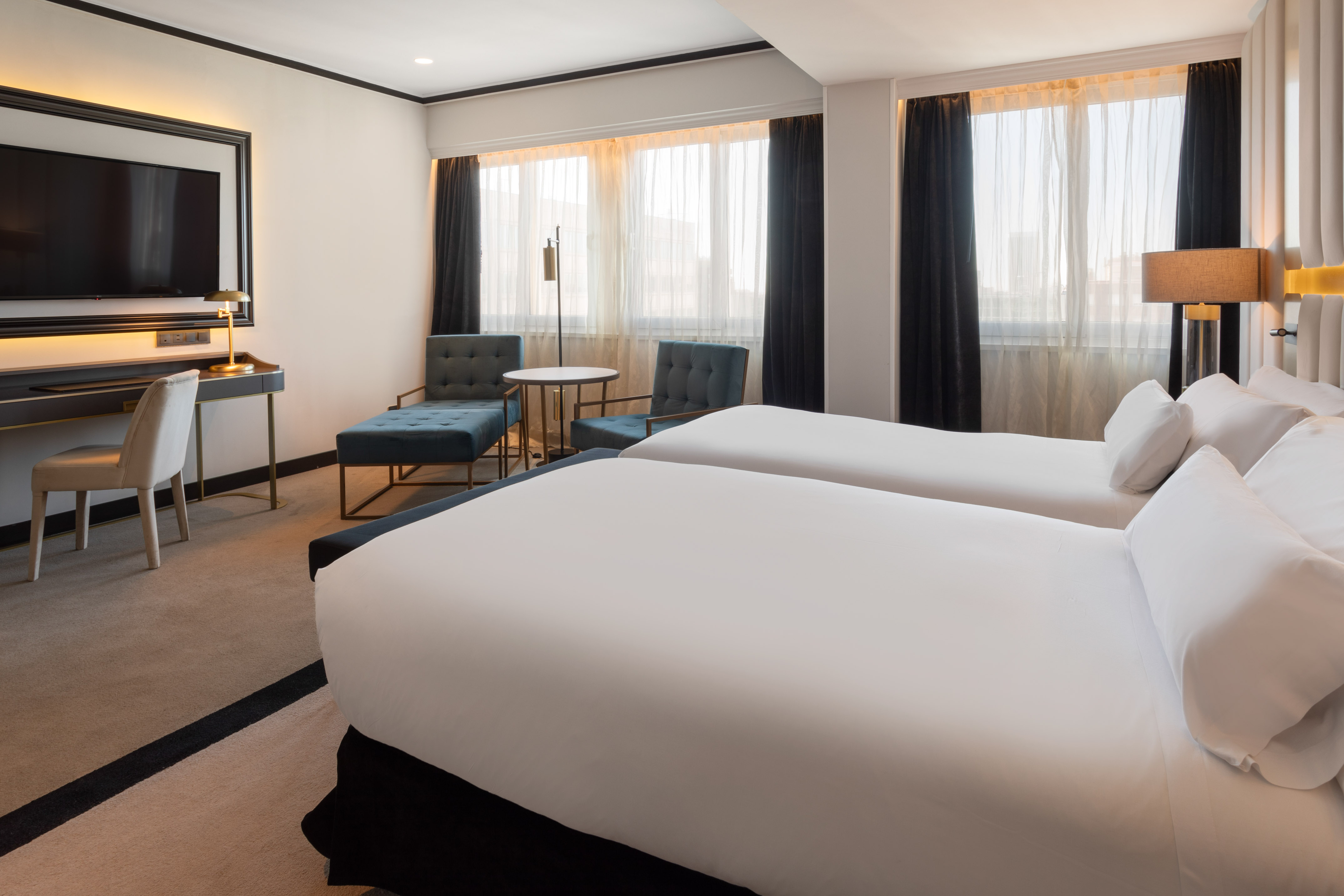 Hotel room with twin beds, TV, desk, and seating by a large window.