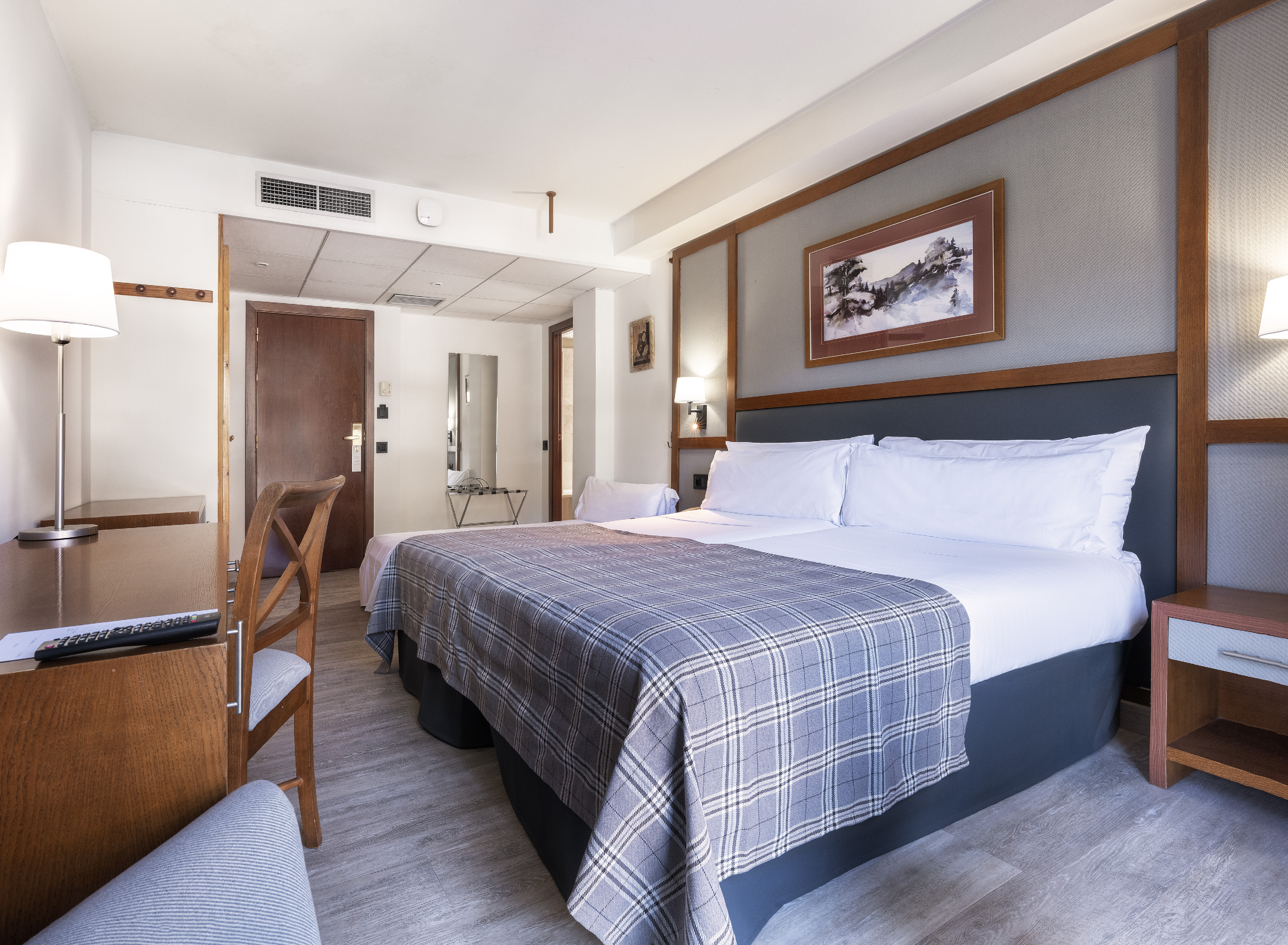 Hotel guest room with large bed, plaid throw, wooden desk, and mountain art.