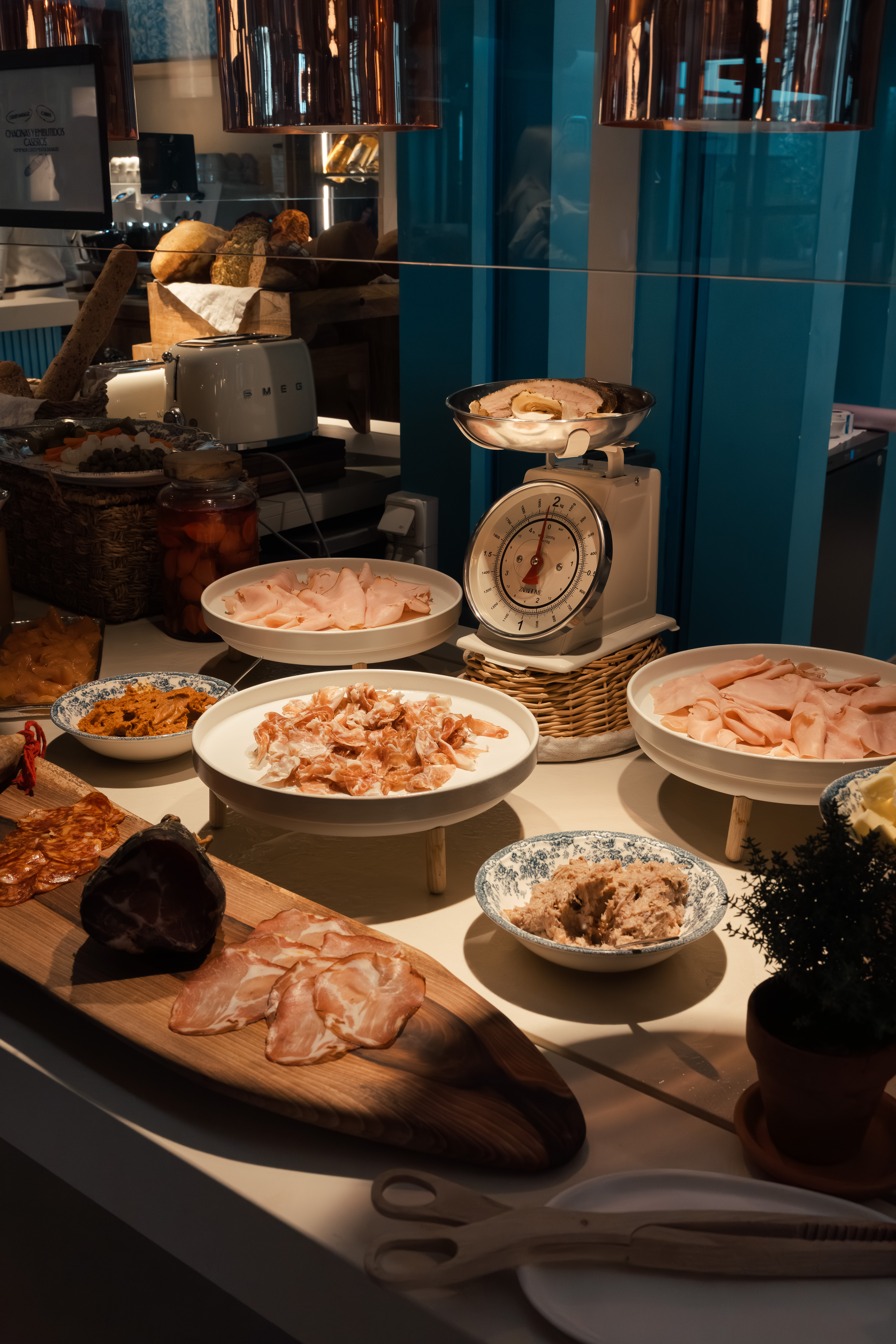 Breakfast buffet with ham, charcuterie, bread, Smeg toaster, and vintage scale.