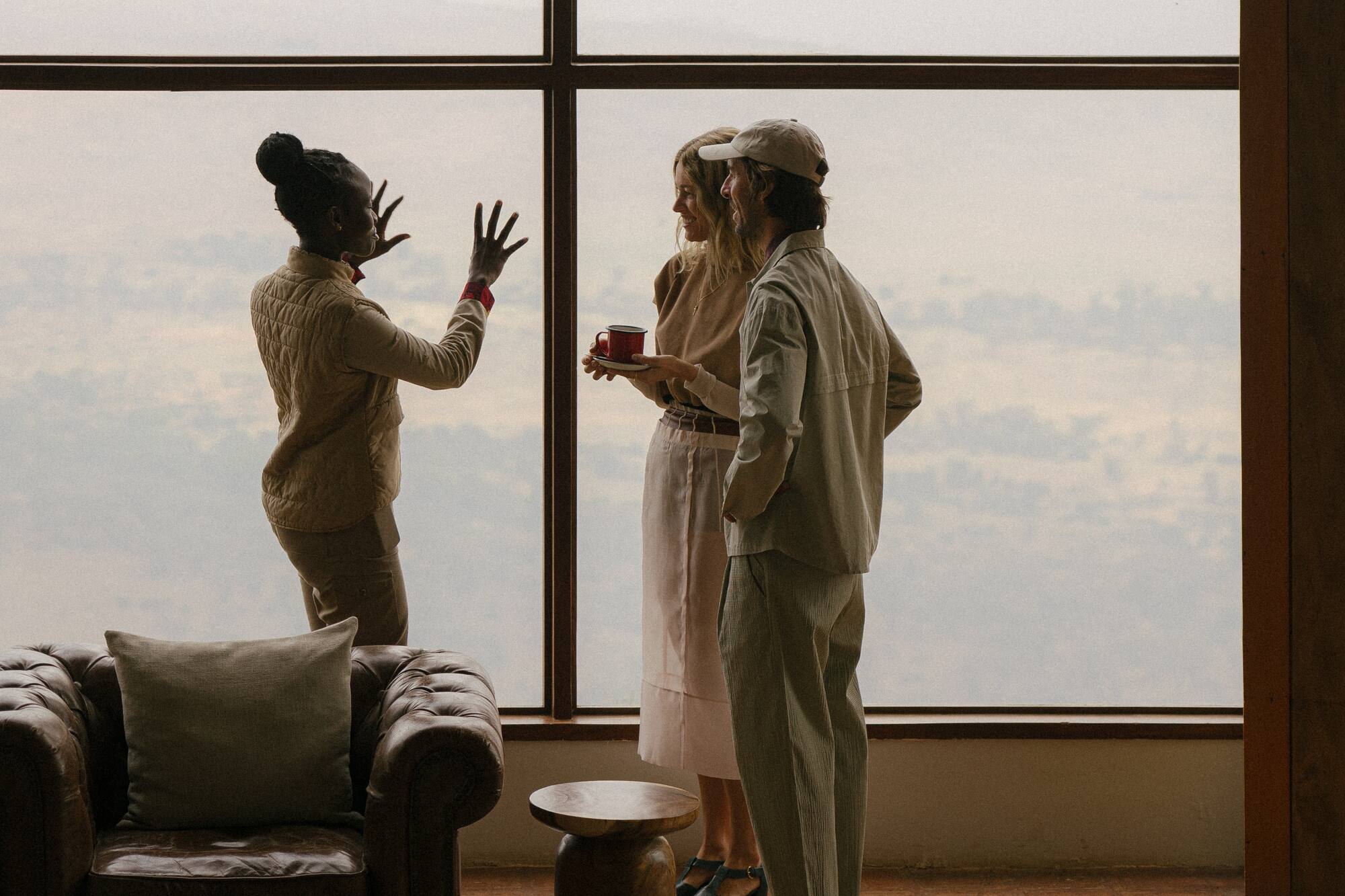Three people conversing by a large window in a lounge, overcast sky view.