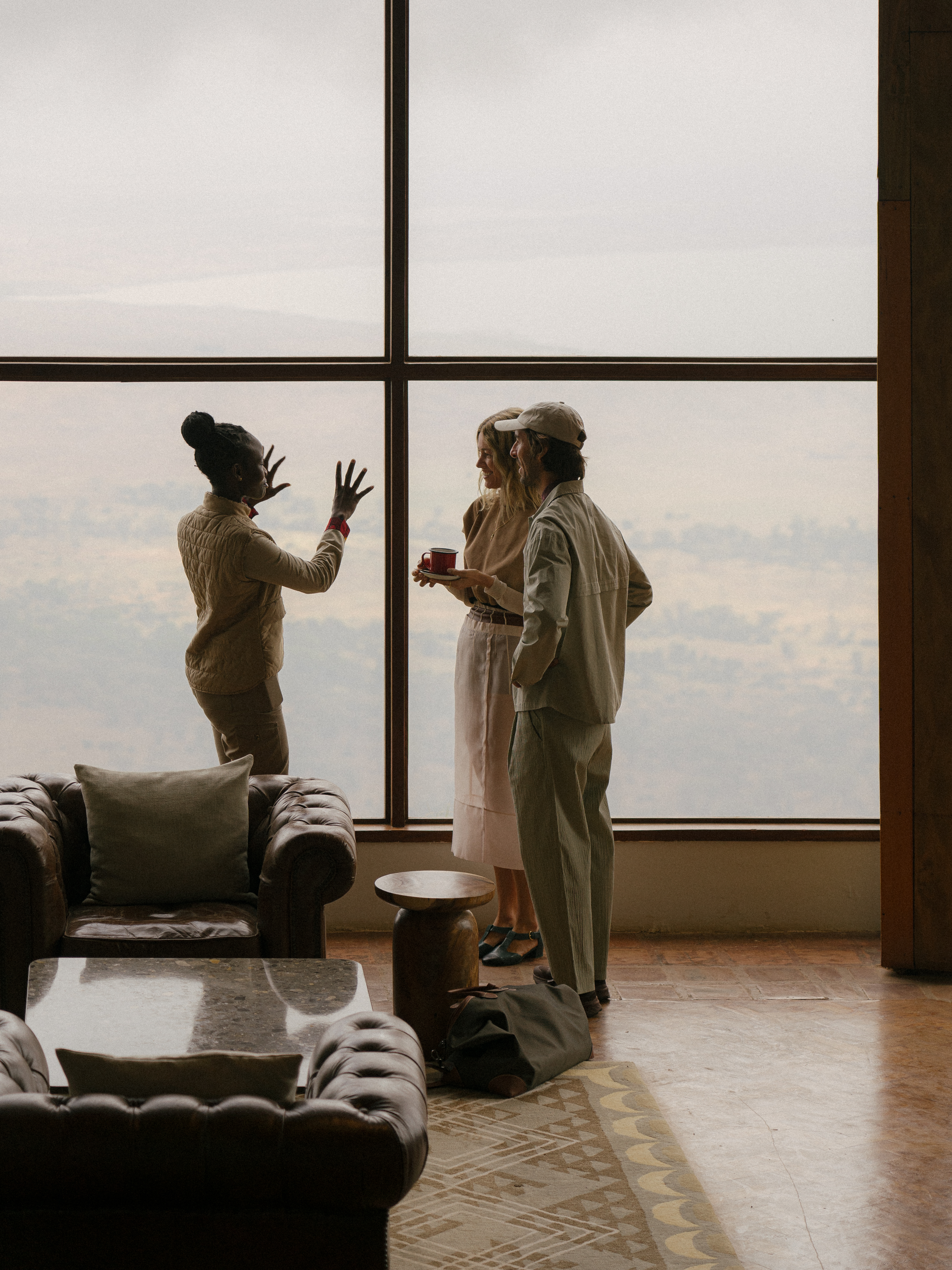 Three people conversing by a large window in a lounge, overcast sky view.