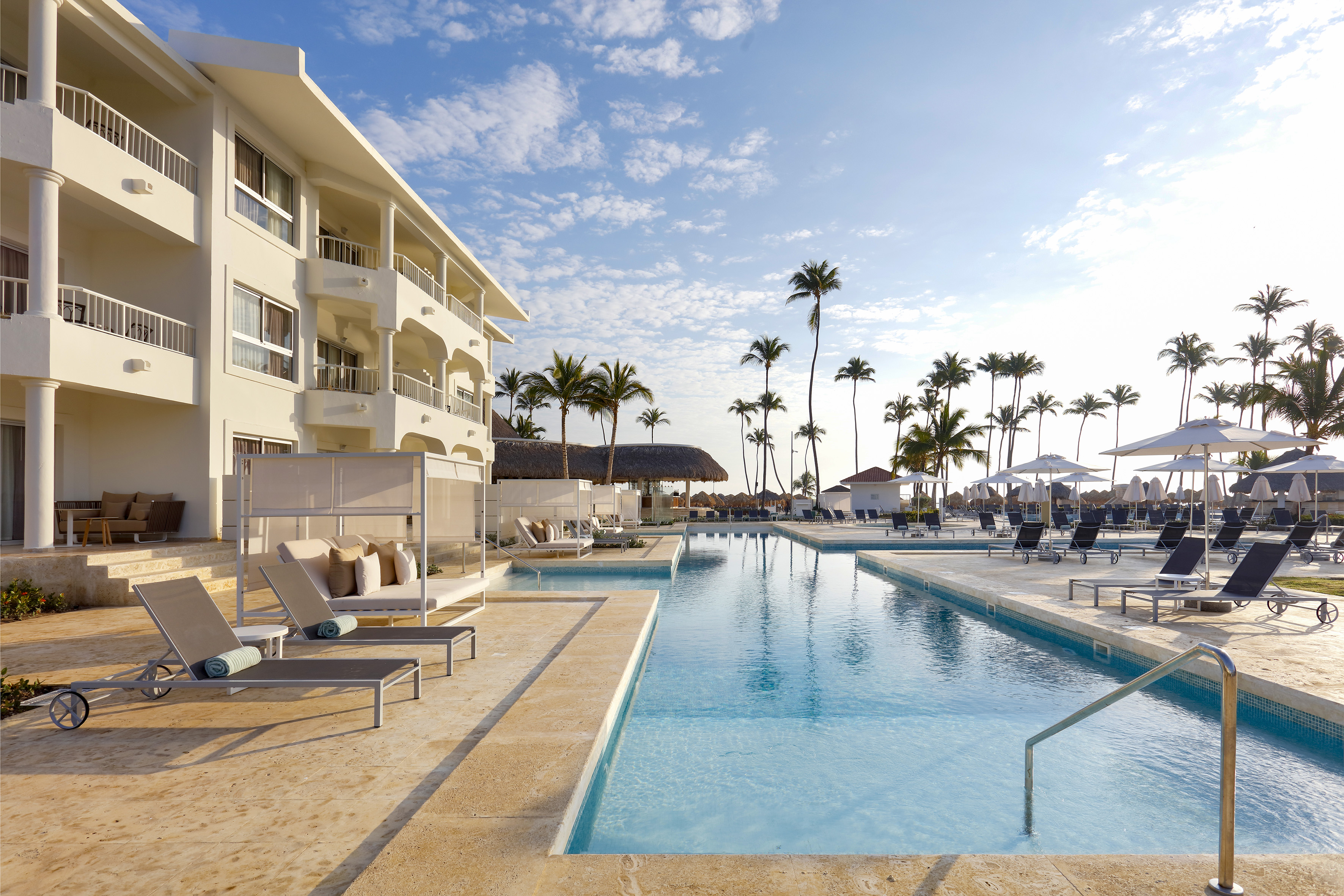 Sunny tropical resort with pool, palm trees, white hotel, and lounge chairs.