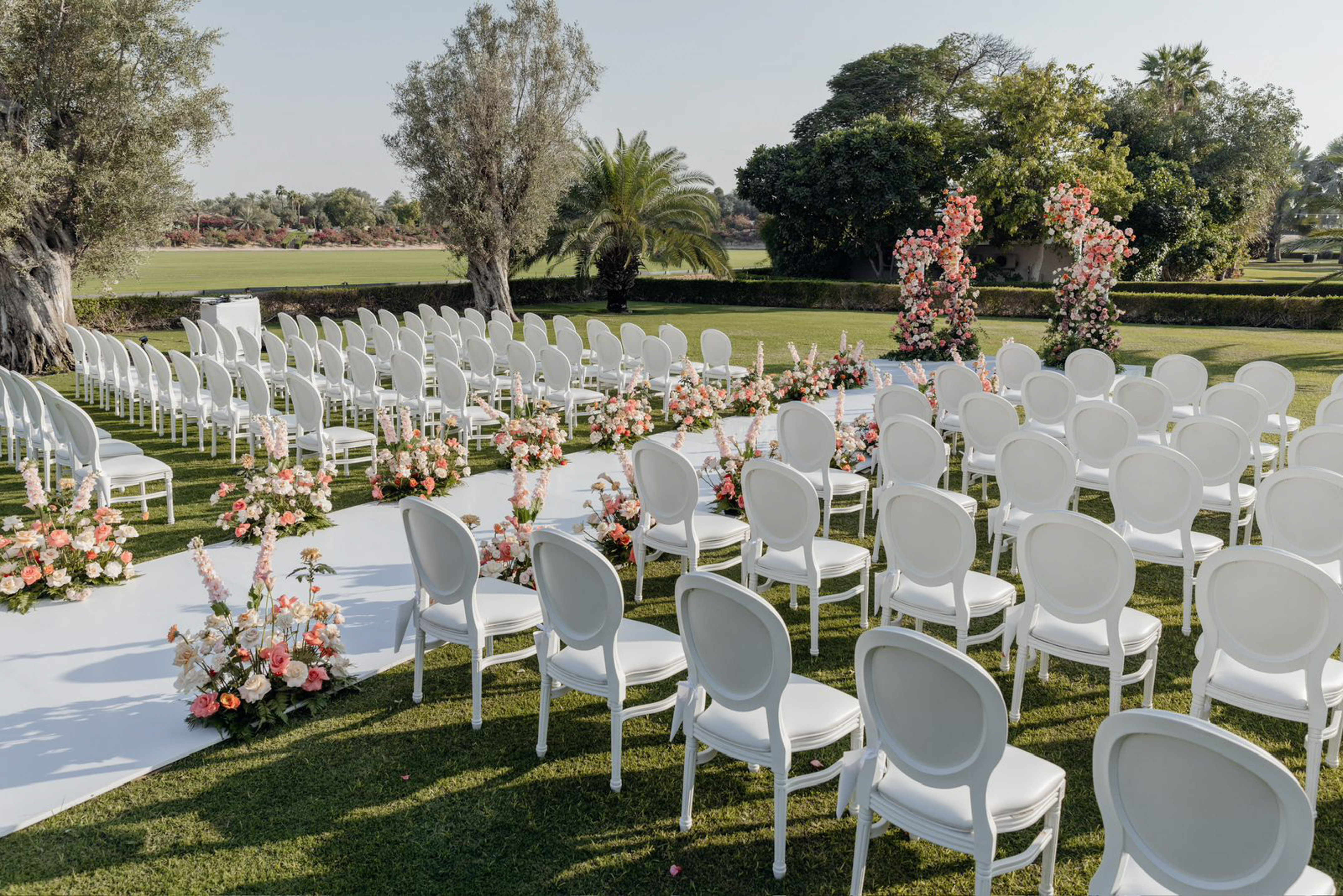 Garden wedding ceremony setup with white chairs, floral aisle, and pink arches.