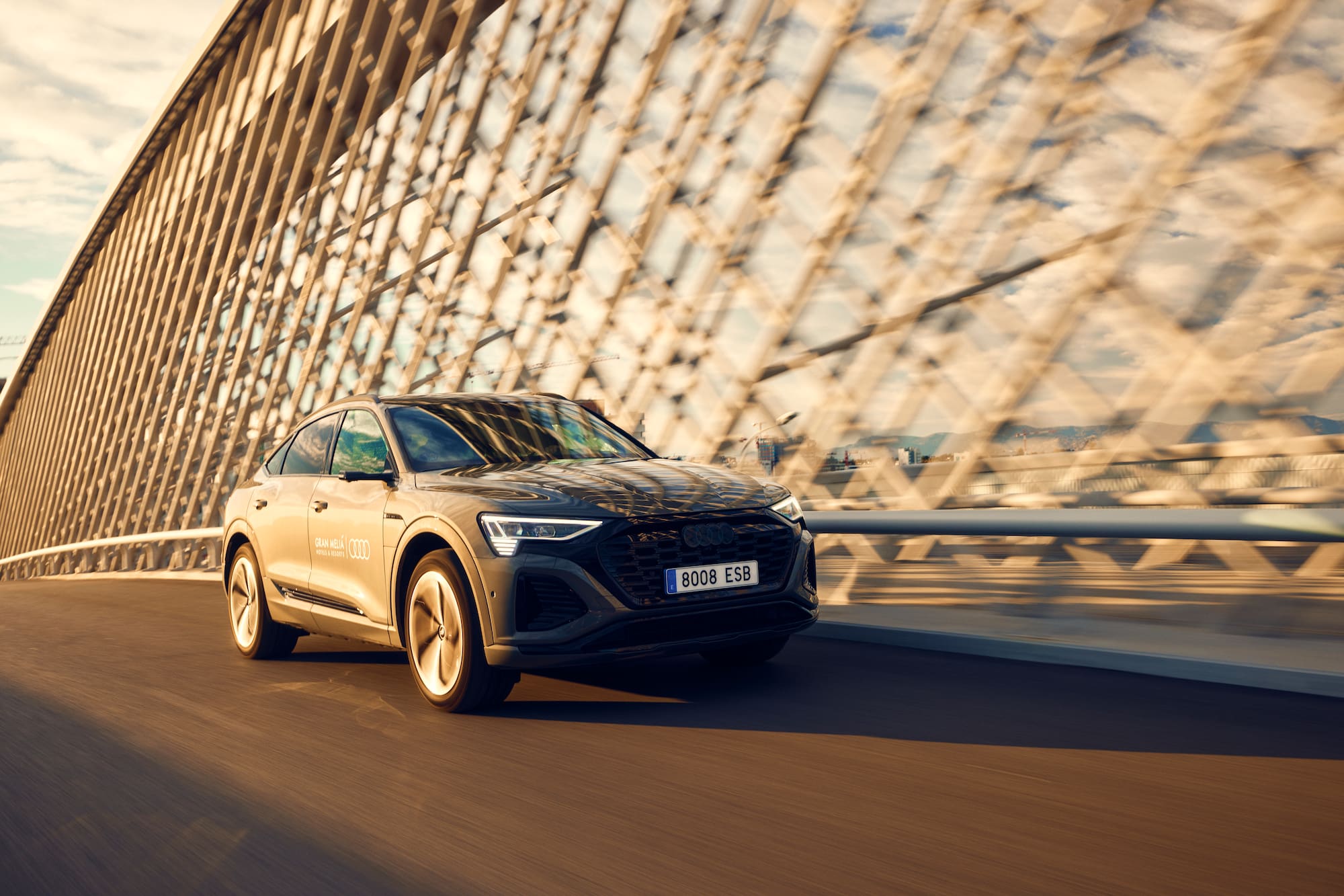 Dark grey Audi e-tron Sportback driving on a modern bridge at sunset.