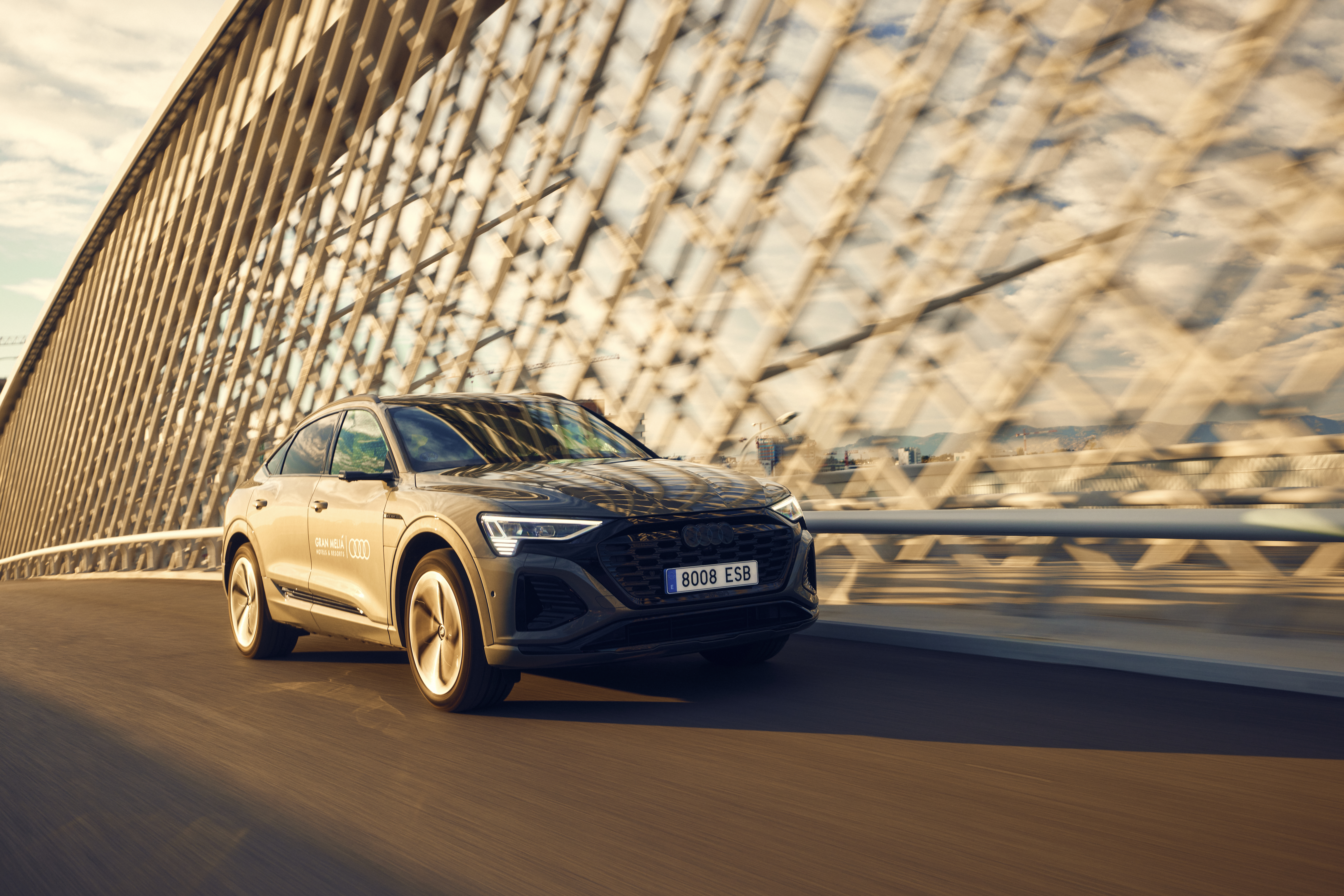 Dark grey Audi e-tron Sportback driving on a modern bridge at sunset.