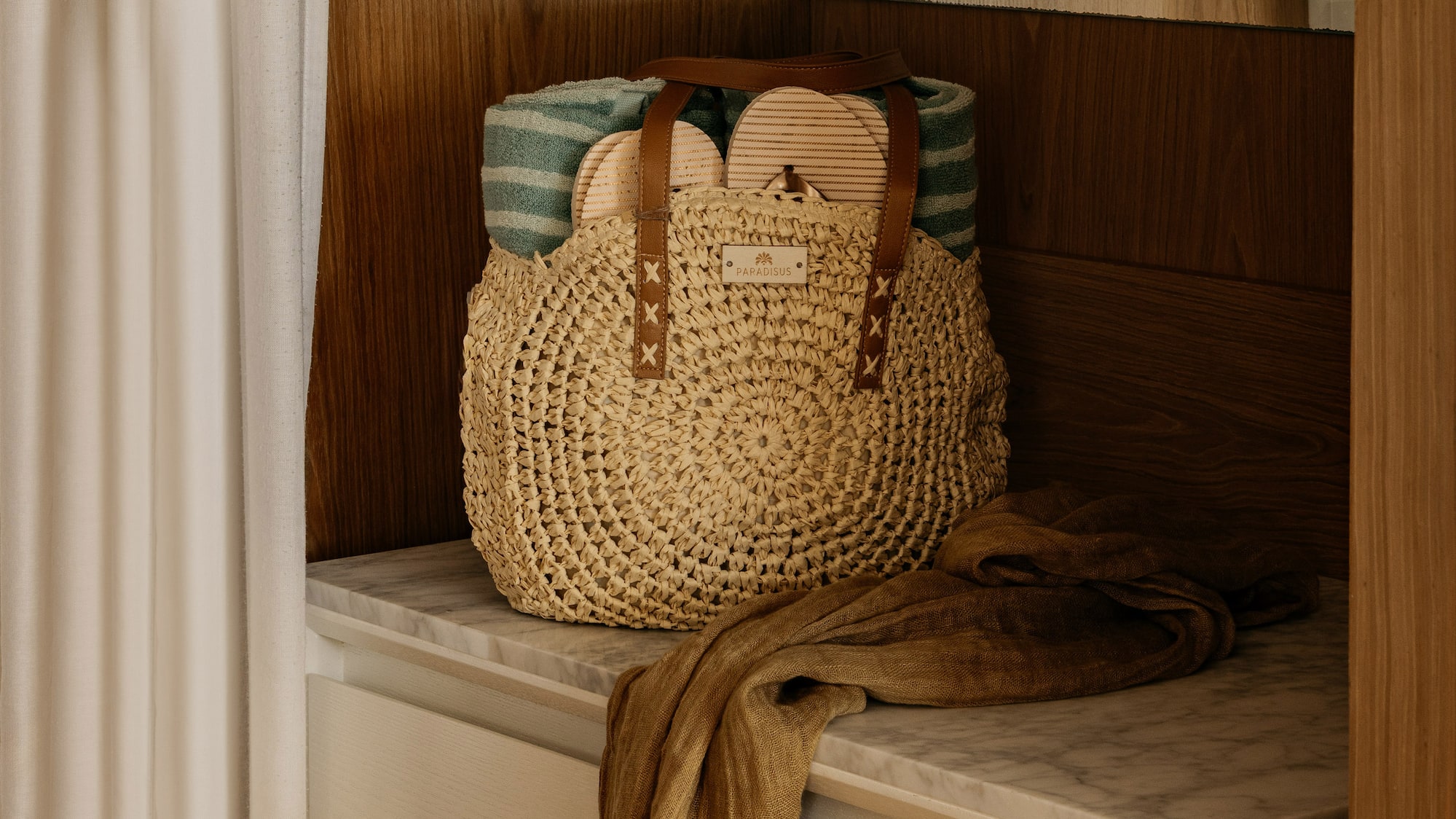 Woven straw beach bag with towel, sandals, and scarf on marble cabinet.