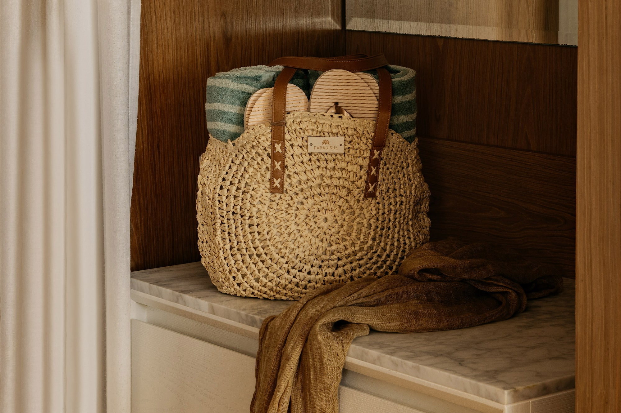Woven straw beach bag with towel, sandals, and scarf on marble cabinet.