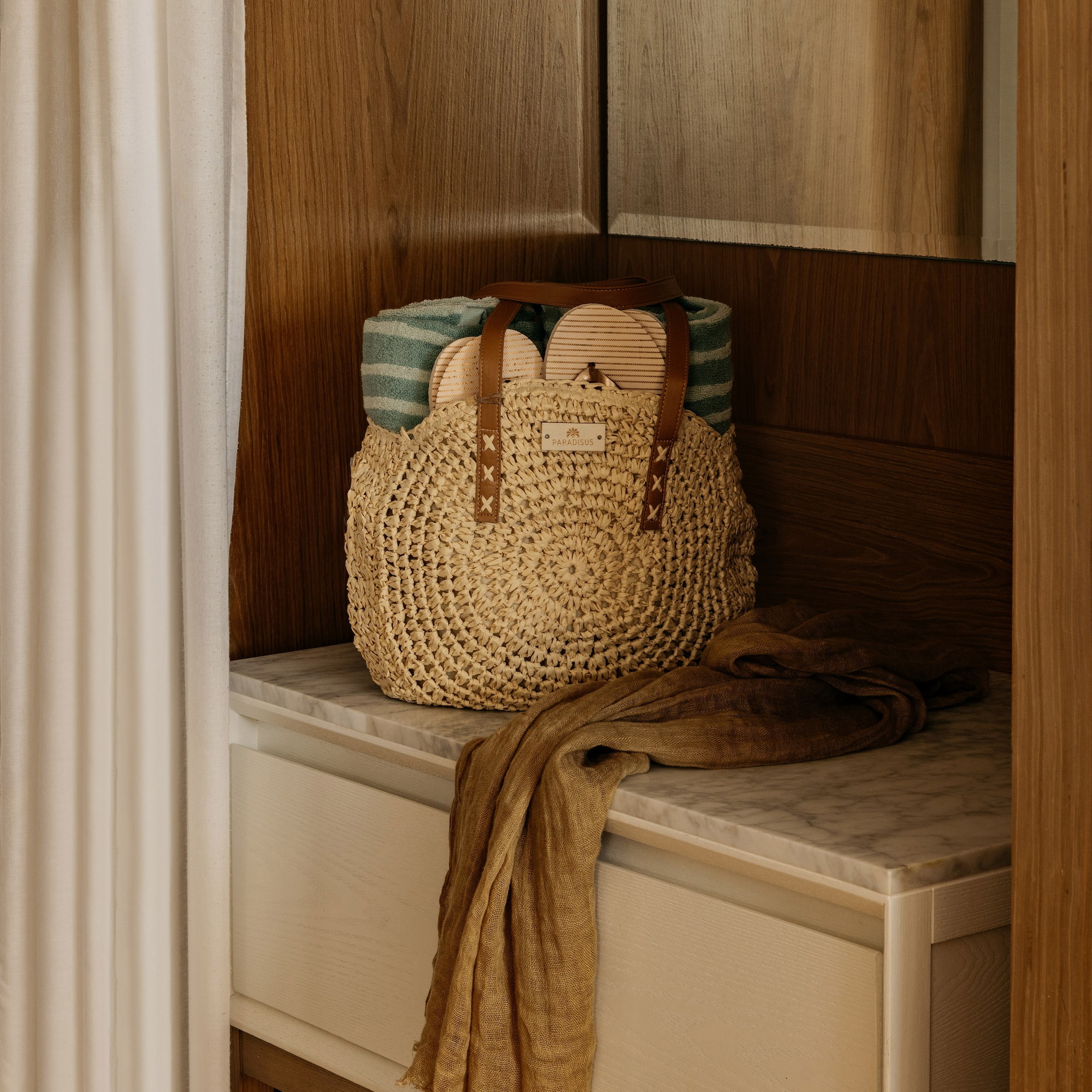 Woven straw beach bag with towel, sandals, and scarf on marble cabinet.