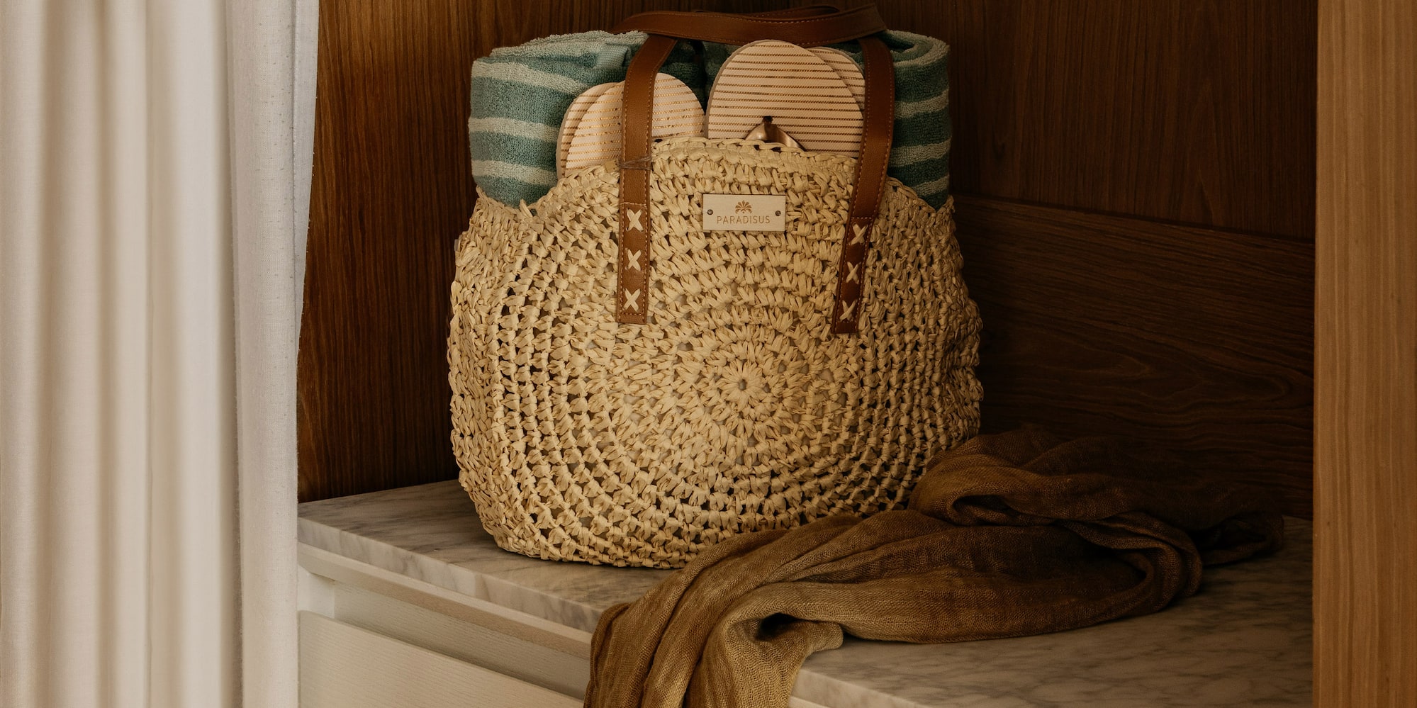 Woven straw beach bag with towel, flip-flops, and sarong on marble counter.