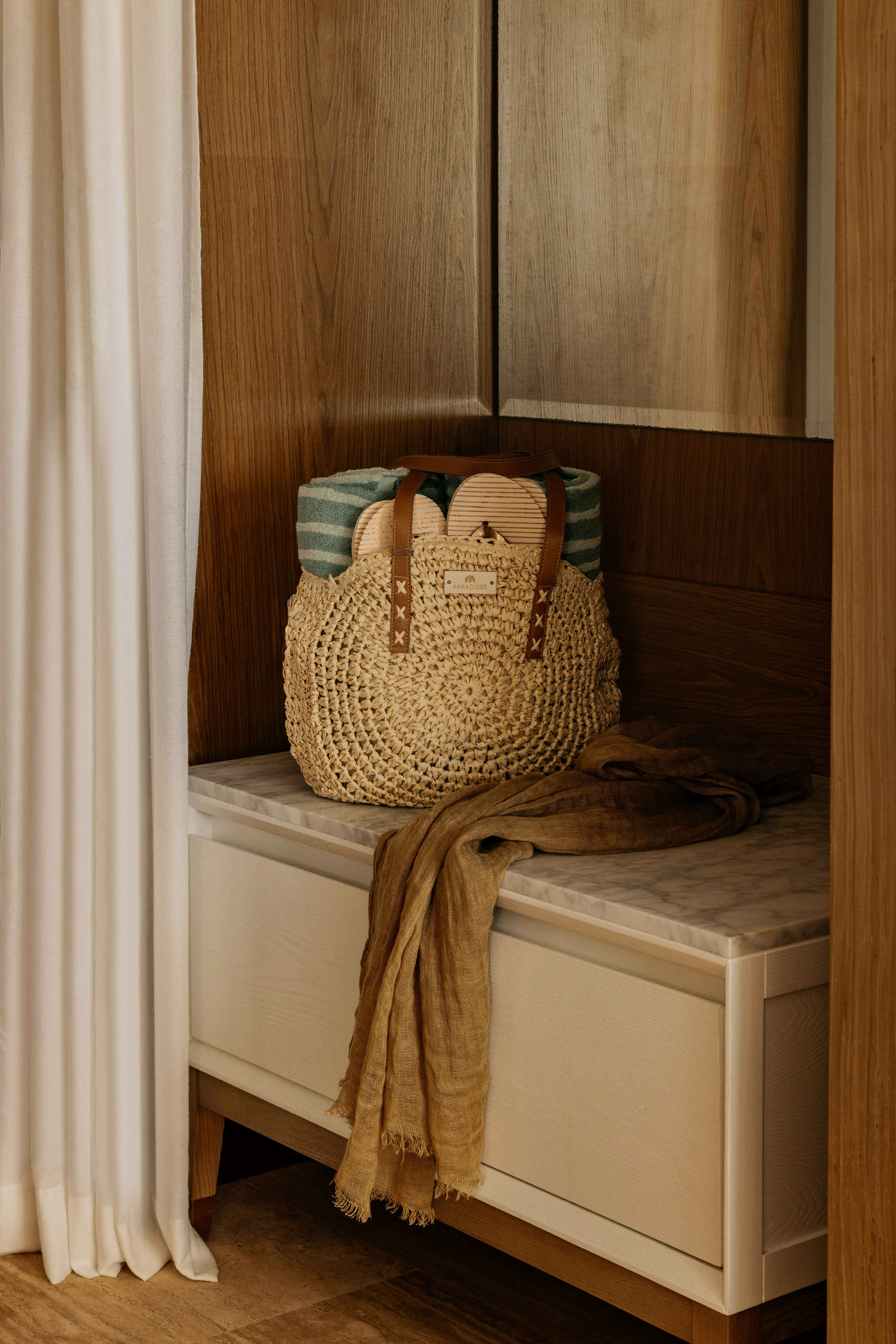 Woven straw beach bag with towel, sandals, and scarf on marble cabinet.