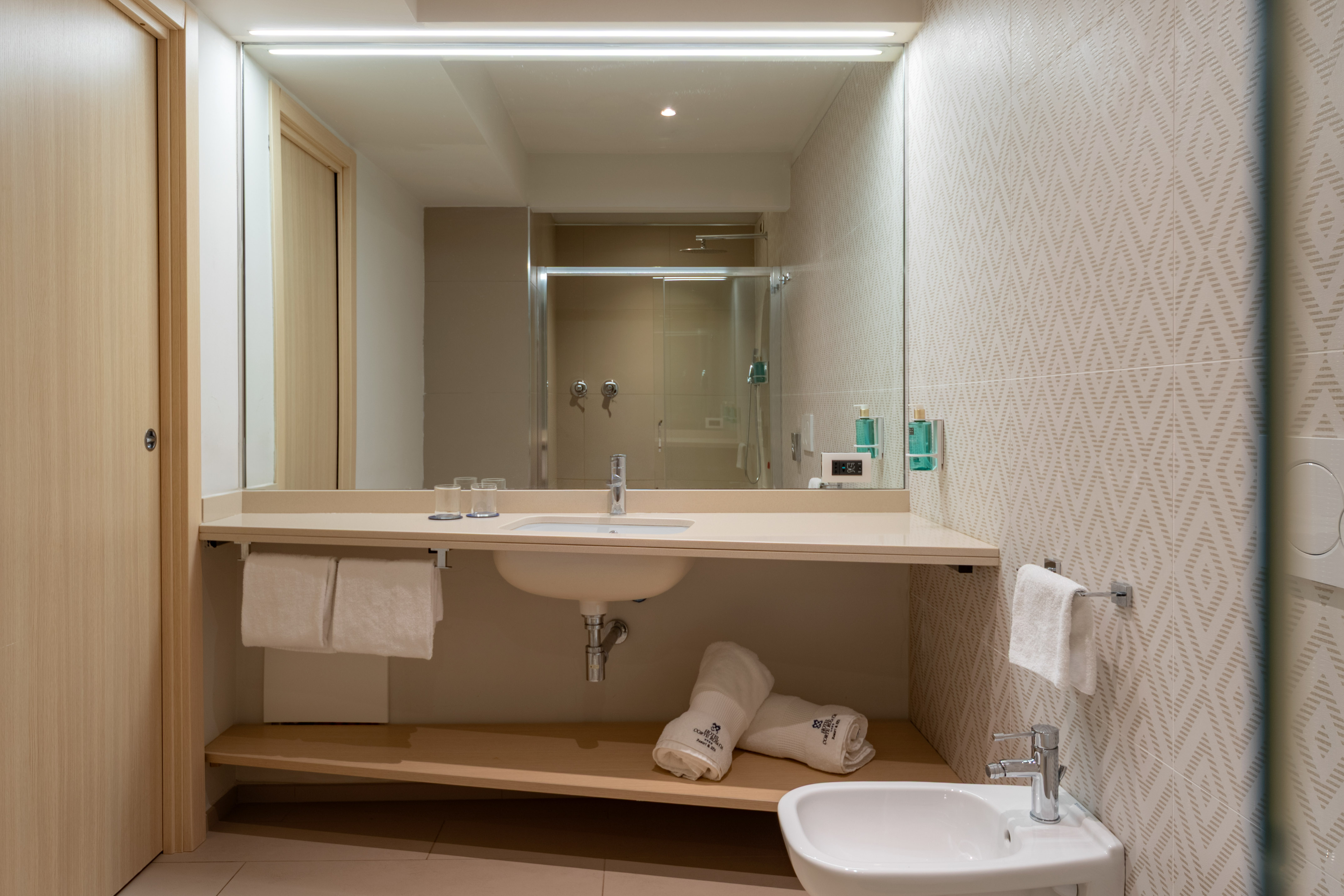 Modern hotel bathroom vanity, sink, mirror reflecting shower, bidet, and towels.