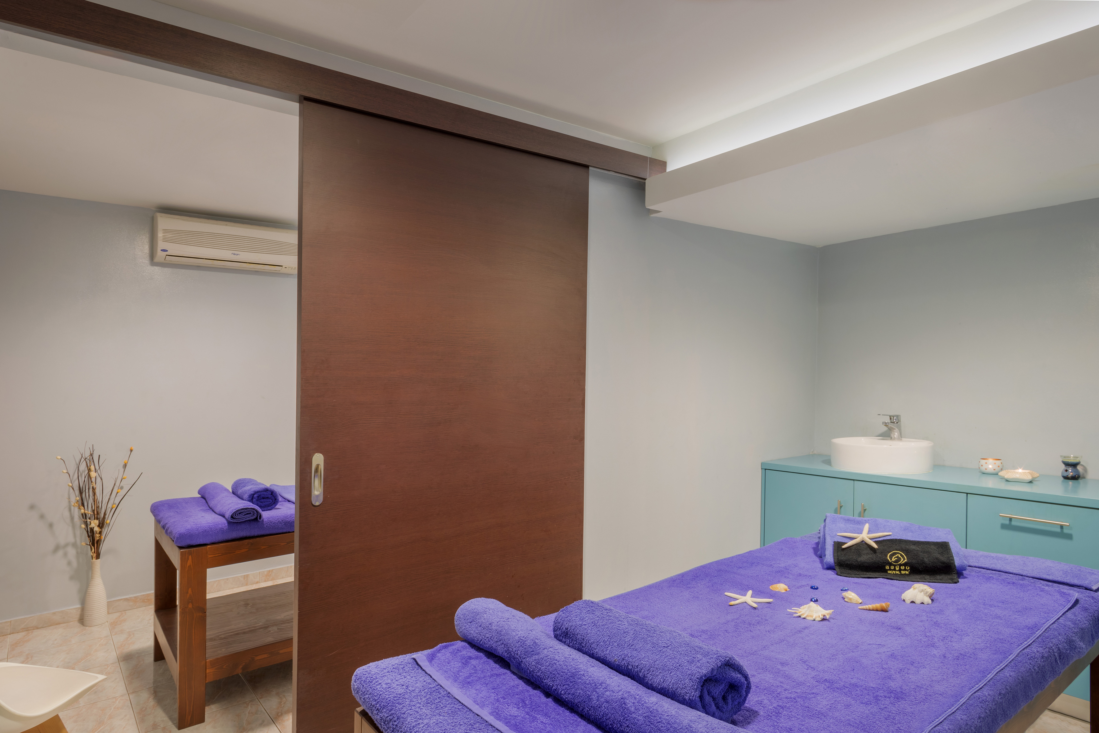 Spa treatment room with two massage beds, purple towels, and seashells.