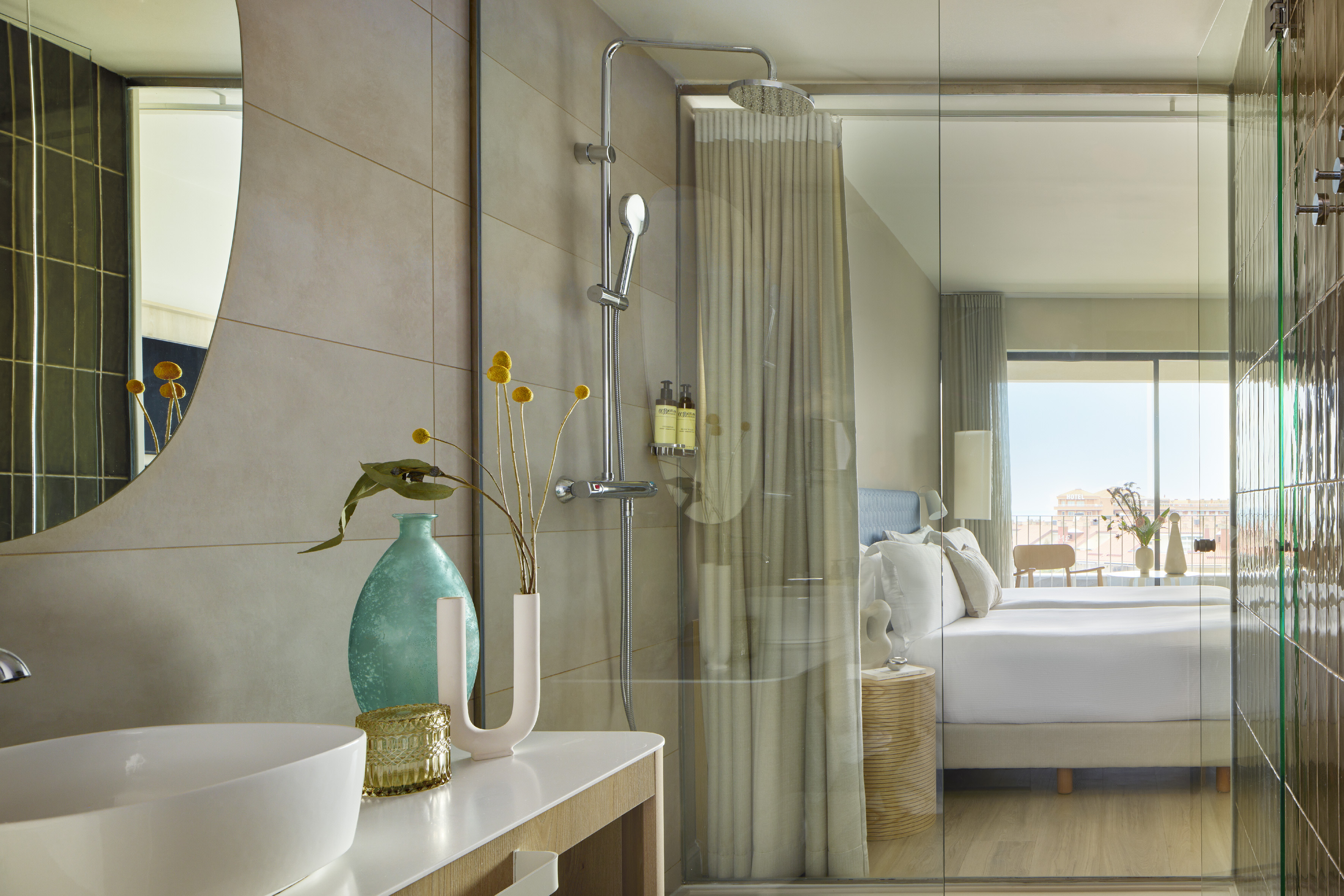 Modern hotel bathroom vanity, glass shower, and bright bedroom.