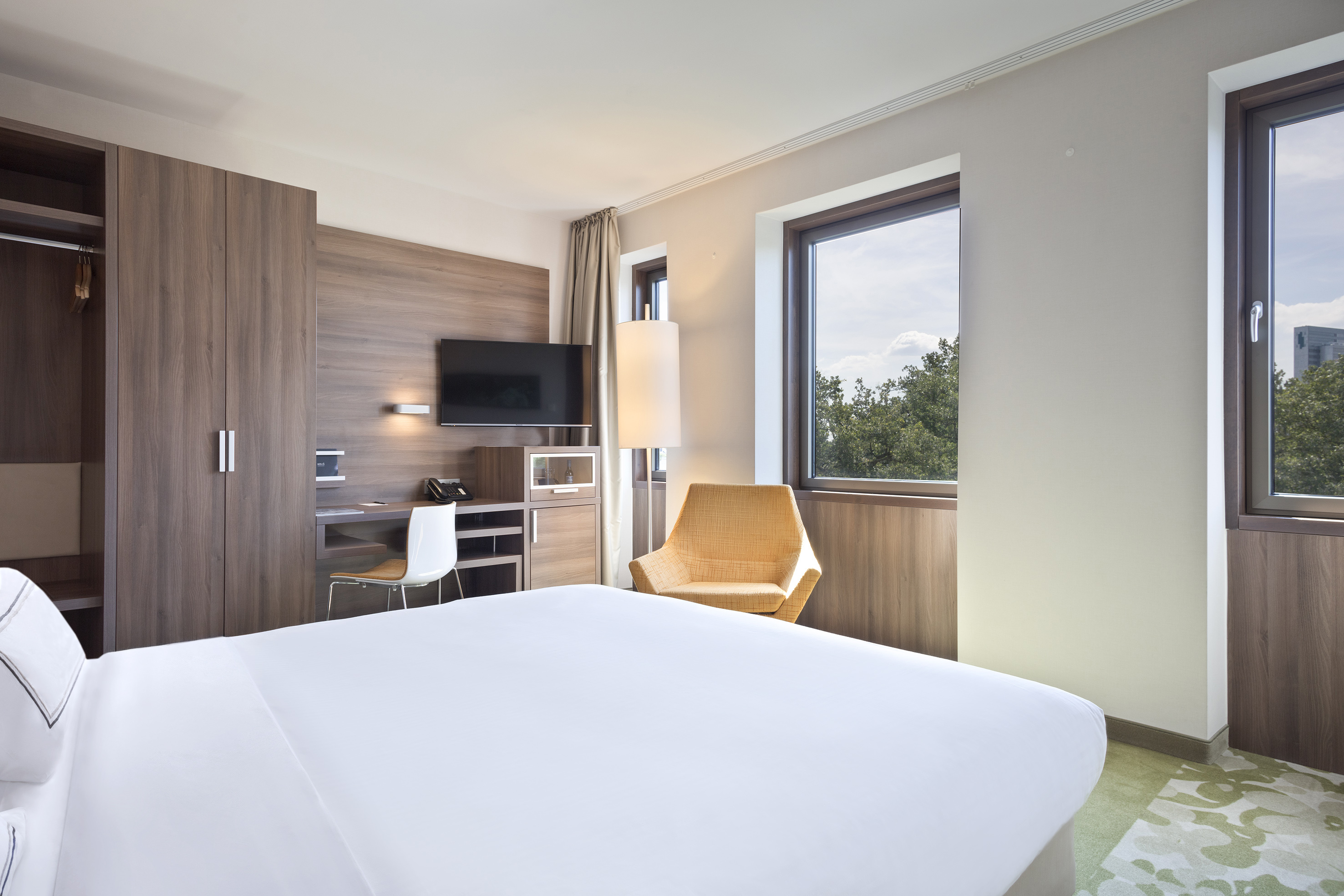 Modern hotel bedroom, white bed, wooden desk, yellow chair, window green view.