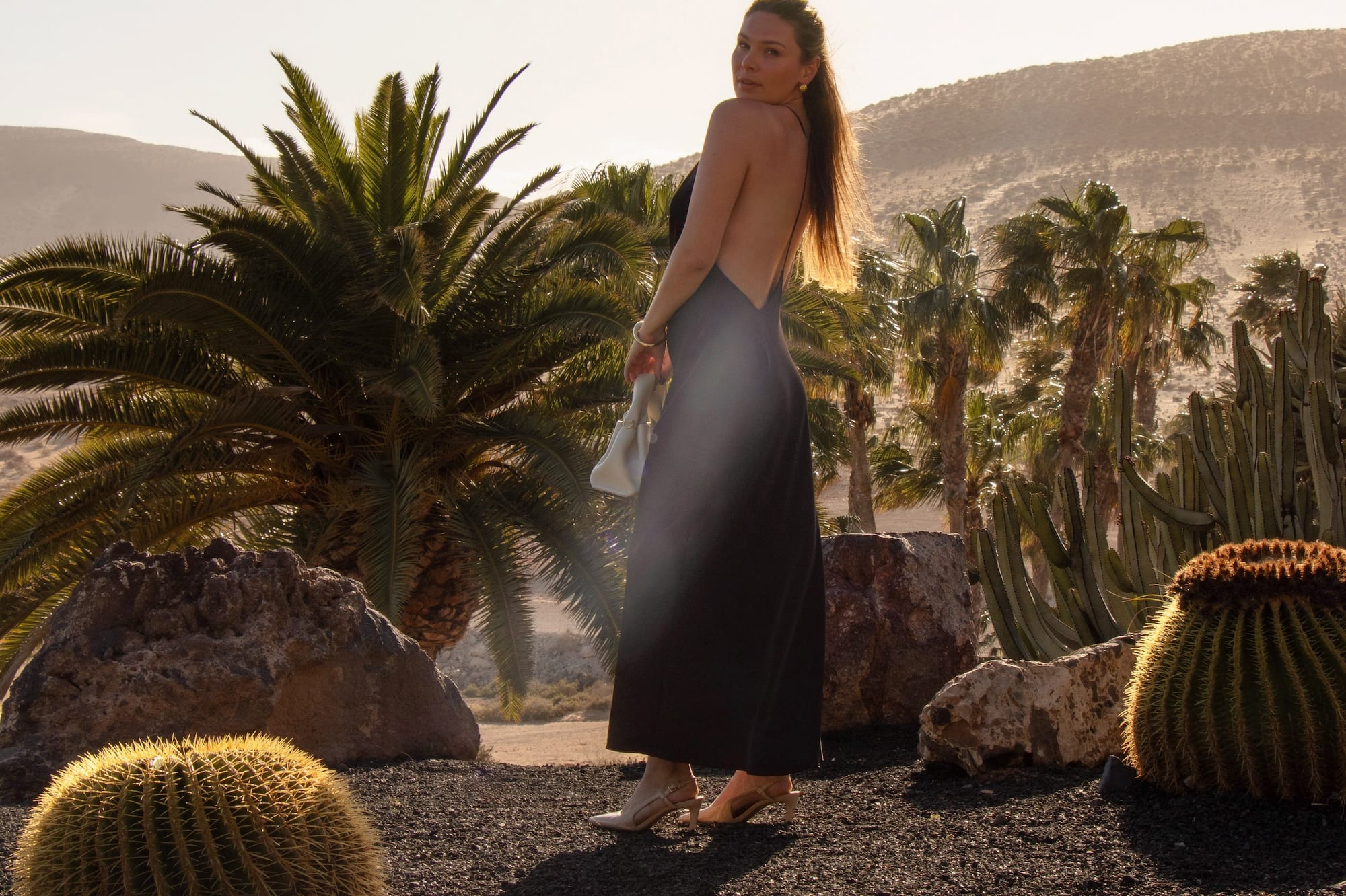 Mujer con vestido negro en jardín desértico con palmeras y cactus al atardecer.