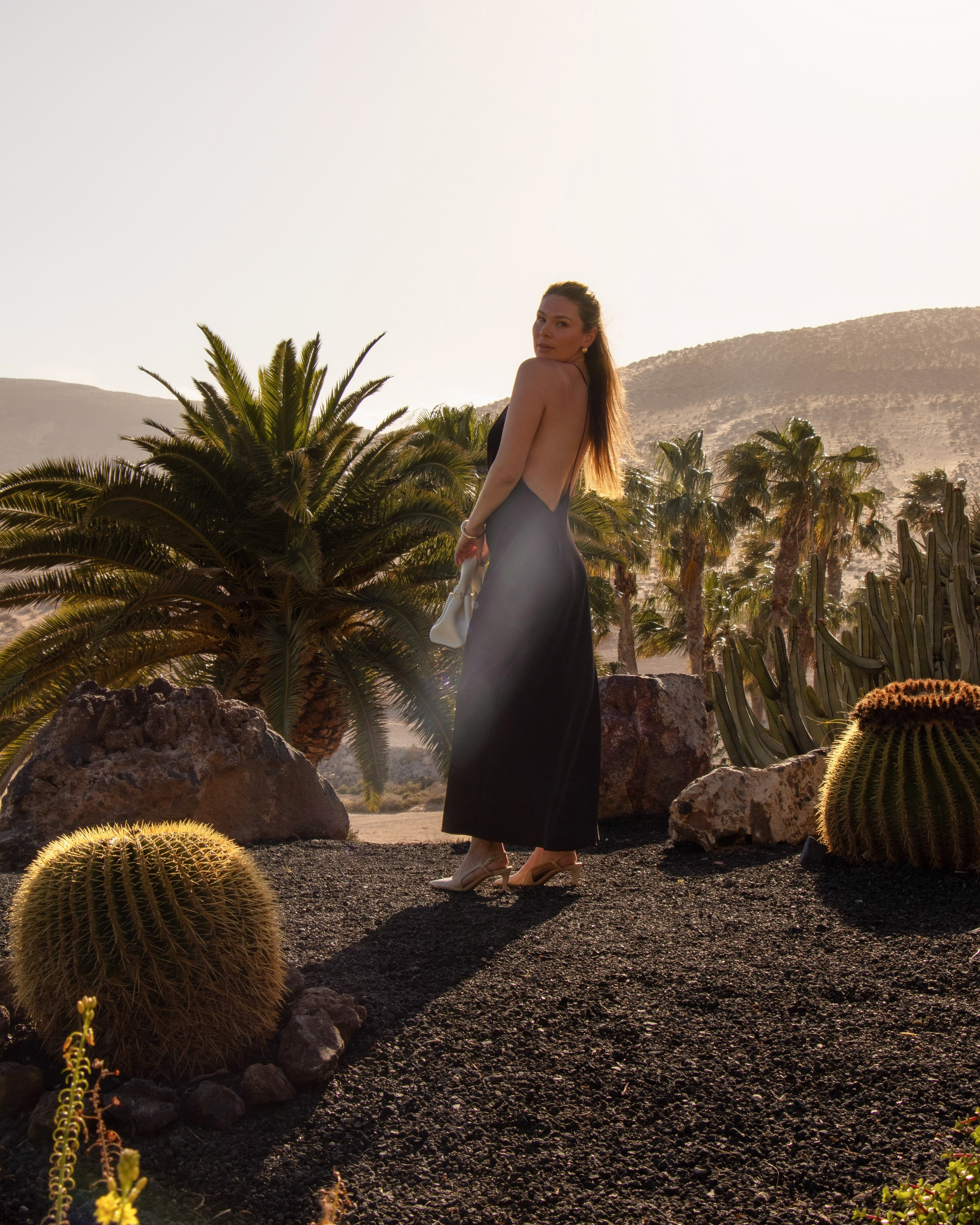 Mujer con vestido negro en jardín desértico con palmeras y cactus al atardecer.