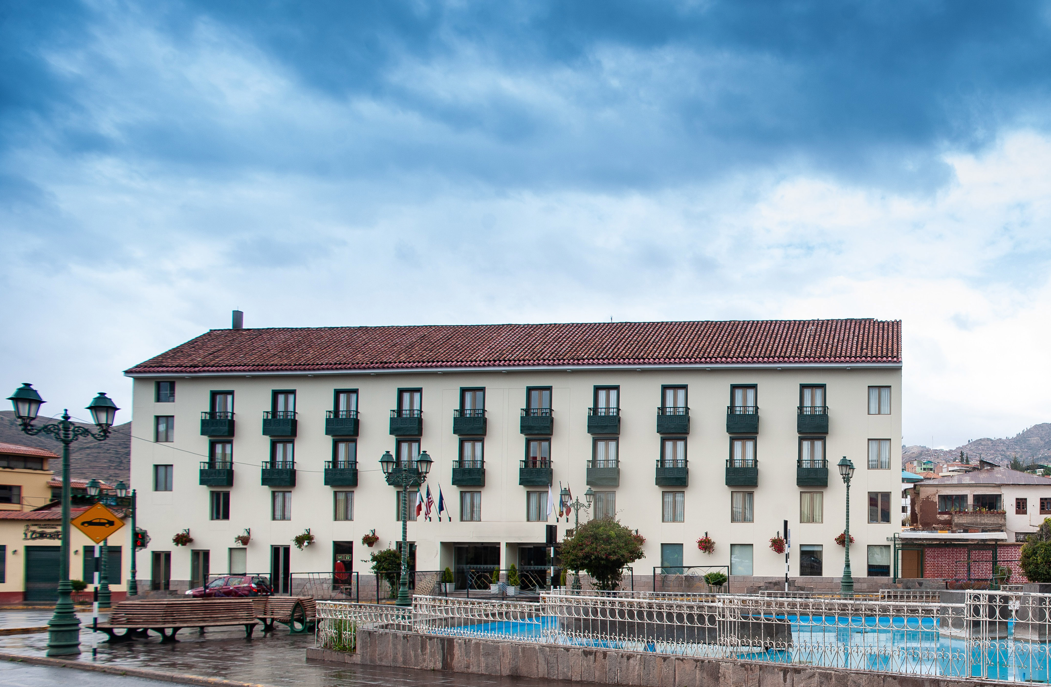 Hotel with red roof, green balconies, and flags, beside a town plaza fountain.