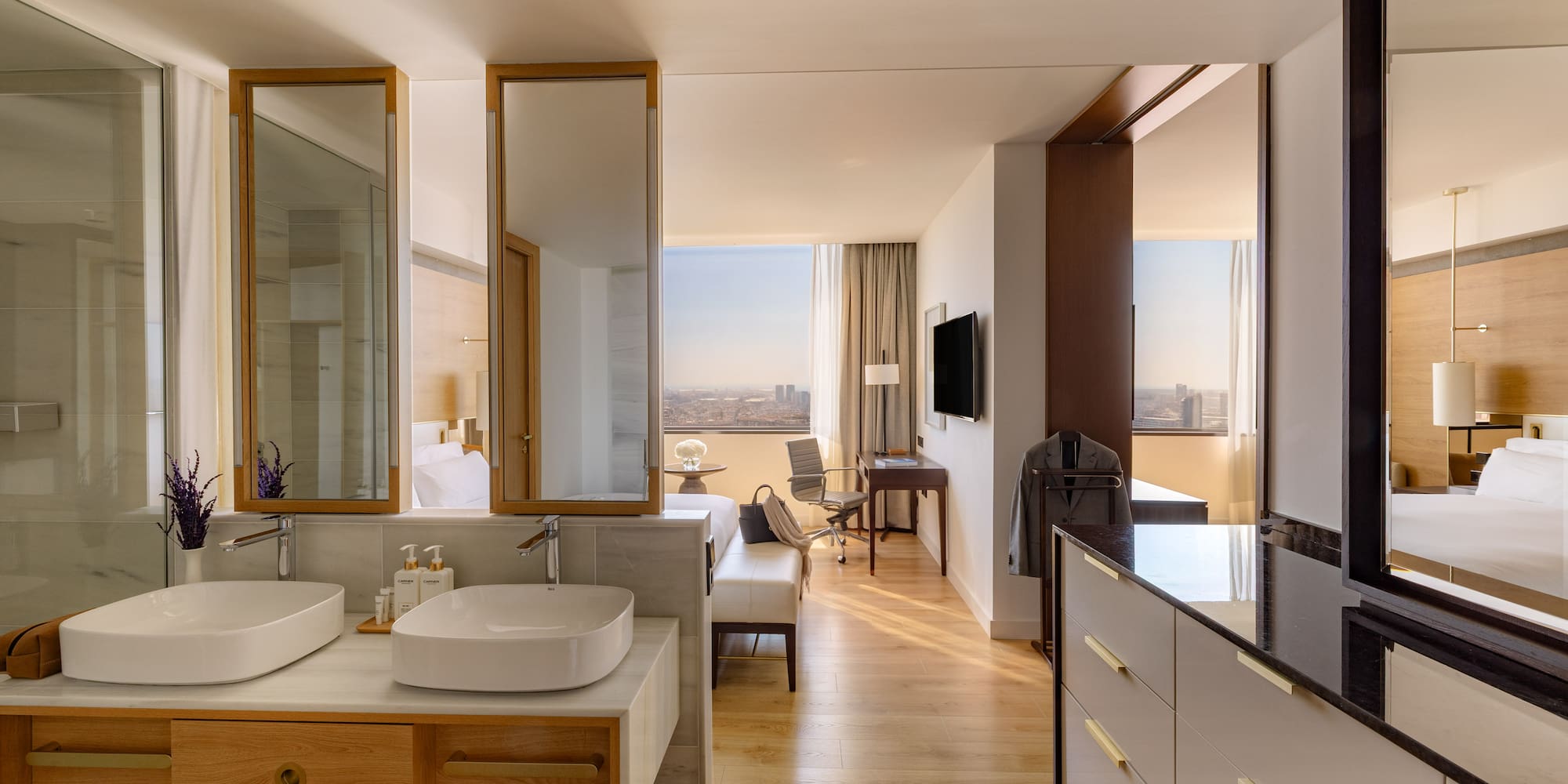 Modern hotel suite with open bathroom, dual sinks, wood floors, and city view.