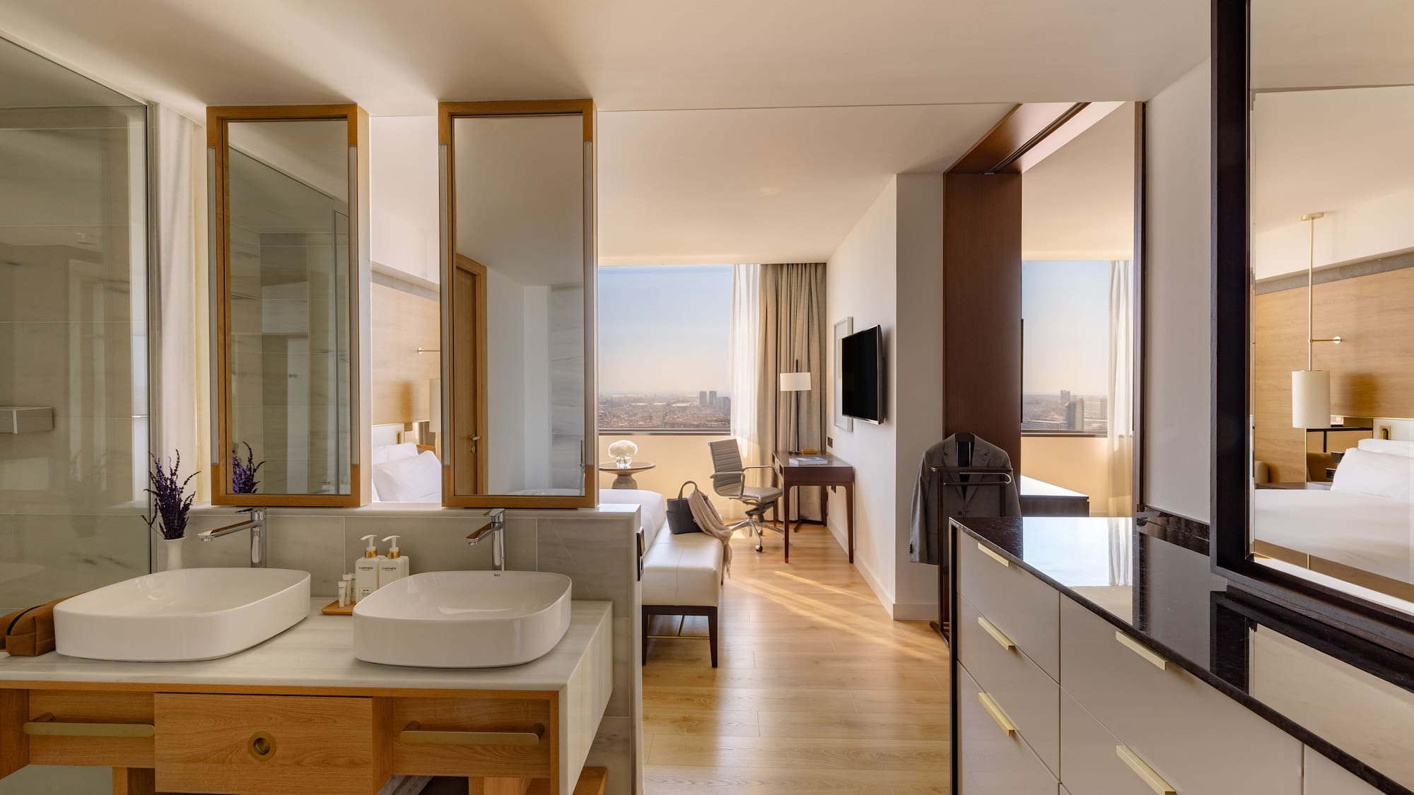 Modern hotel suite with open bathroom, dual sinks, wood floors, and city view.