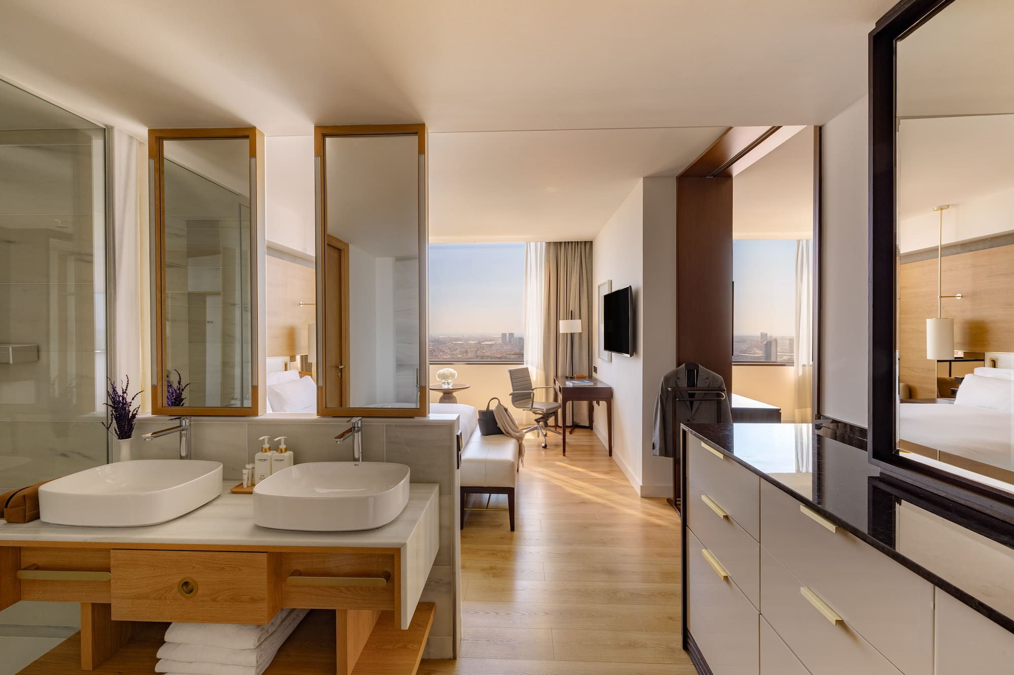 Modern hotel suite with open bathroom, dual sinks, wood floors, and city view.