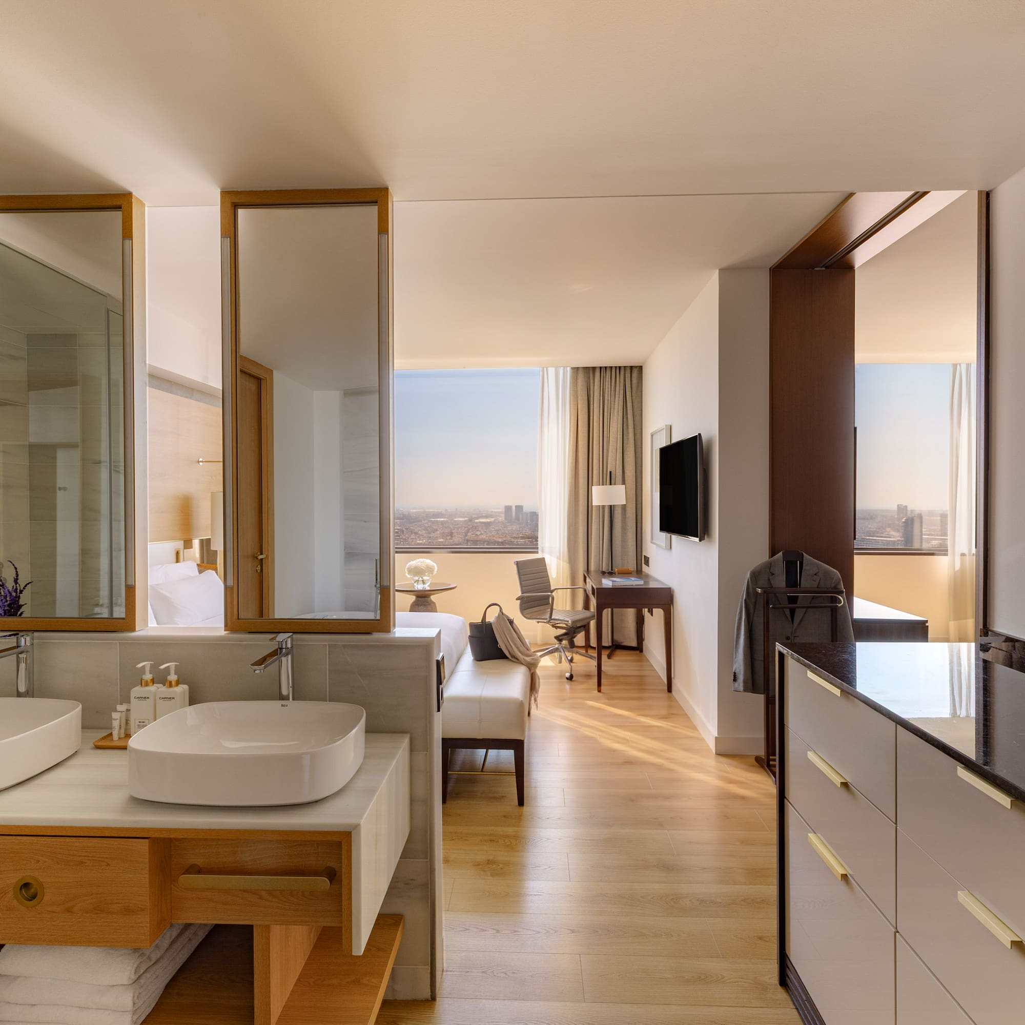 Modern hotel suite with open bathroom, dual sinks, wood floors, and city view.